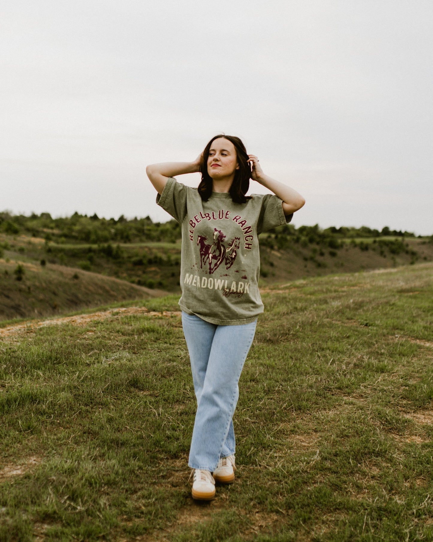 Rebel Blue Ranch Mustangs Tee. A woman wears a mineral wash olive green boxy oversized Rebel Blue Ranch tee with a horses design based on the cowboy romance book series by Lyla Sage that includes Done and Dusted, Swift and Saddled, Lost and Lassoed, and Wild and Wrangled. Meadowlark bookish merch. Lyla Sage merch.