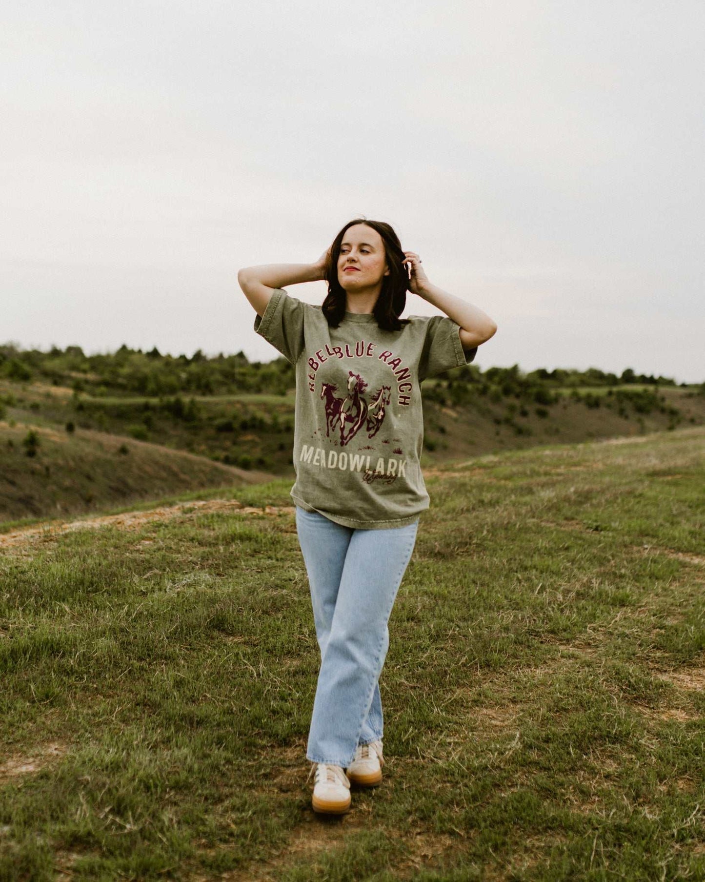 Rebel Blue Ranch Mustangs Tee. A woman wears a mineral wash olive green boxy oversized Rebel Blue Ranch tee with a horses design based on the cowboy romance book series by Lyla Sage that includes Done and Dusted, Swift and Saddled, Lost and Lassoed, and Wild and Wrangled. Meadowlark bookish merch. Lyla Sage merch.