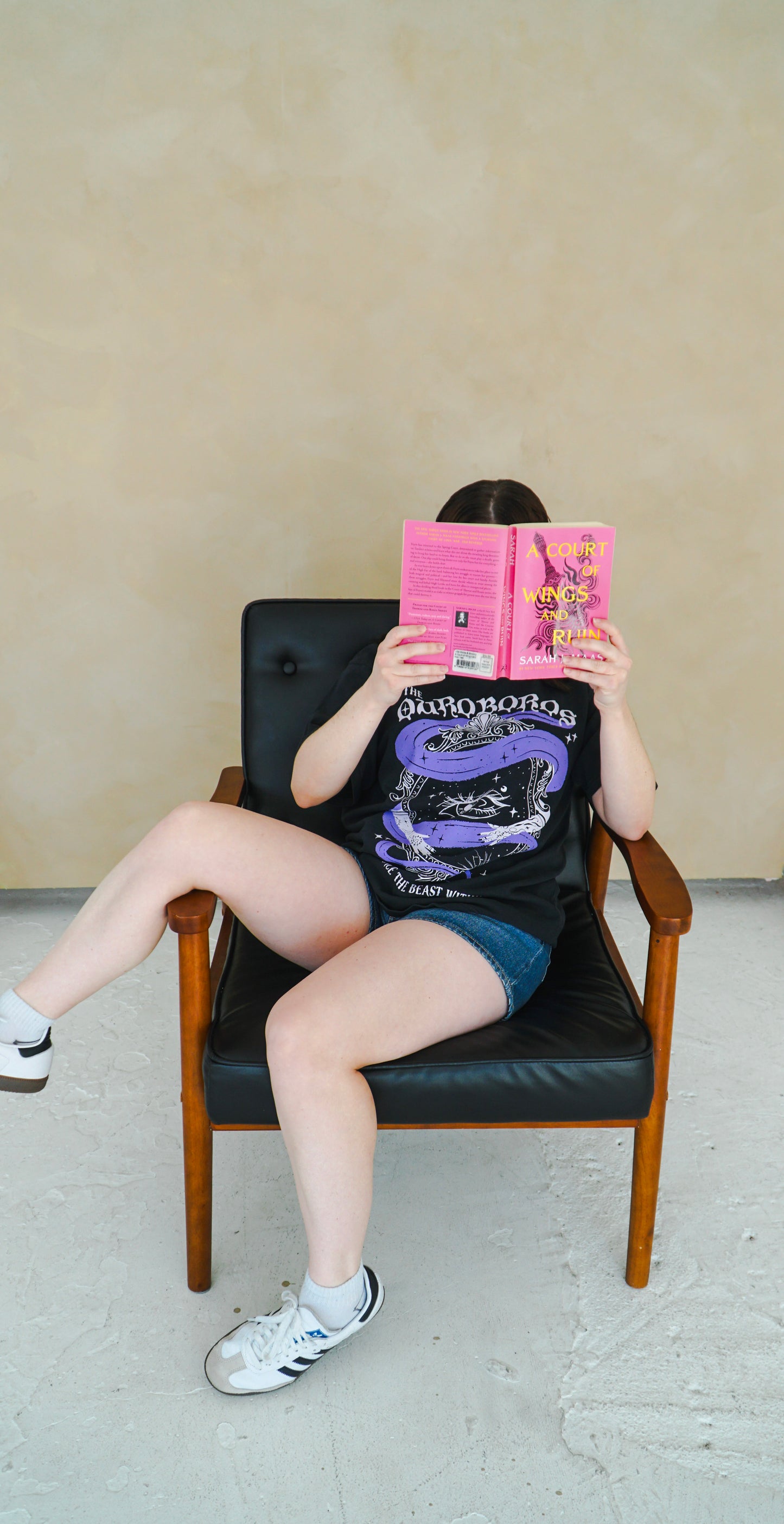 Person sitting on a chair holding a pink book with a plain background. She is wearing a black graphic tee with 'The Ourororos' design featuring an eye, mirror, and purple mist. Inspired by the A Court of Thorns of Roses book series by Sarah J. Maas.
