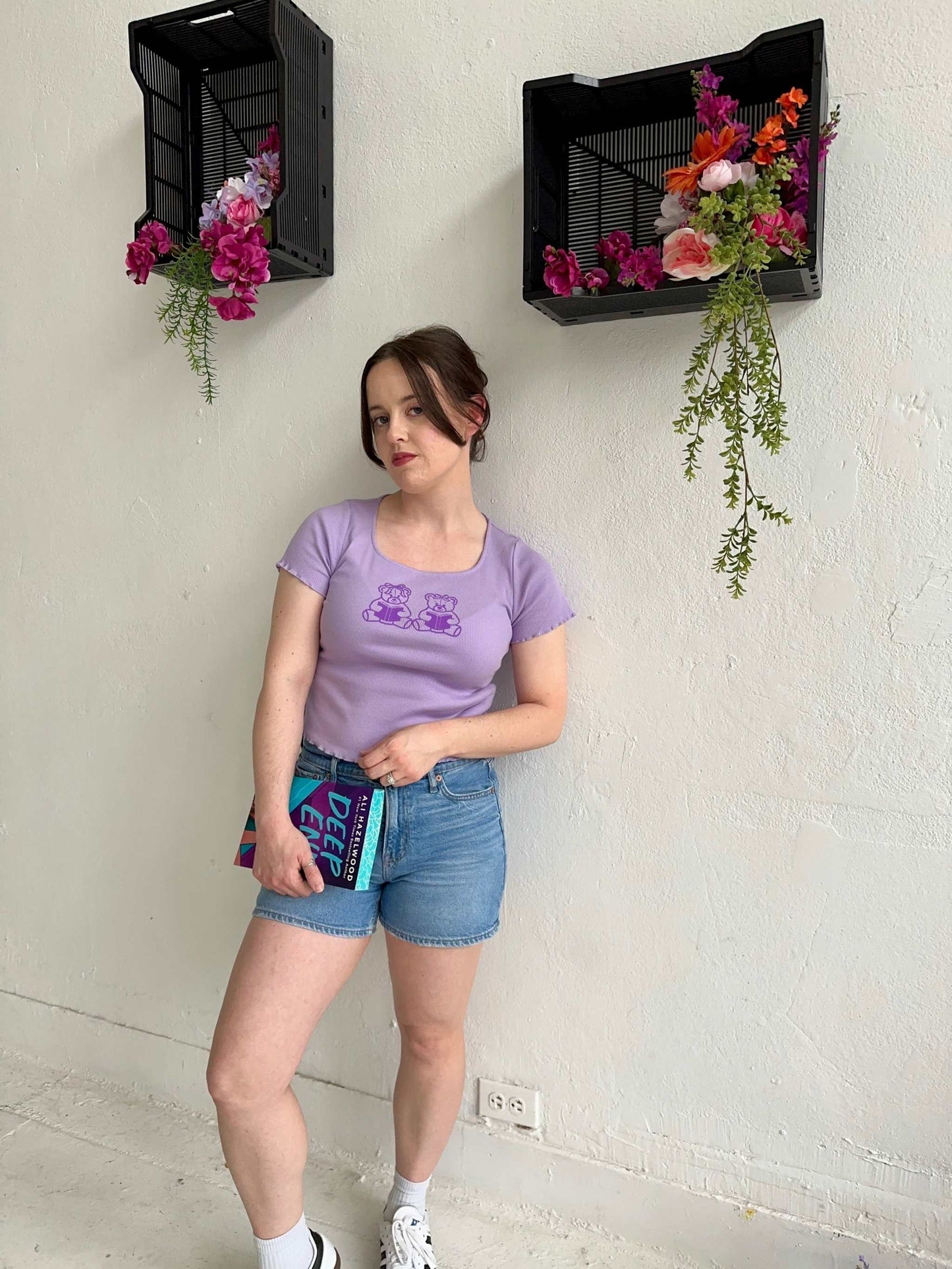 The Book Bears Baby Tee features a design with two teddy bears each holding a book. The t-shirt is a light purple color with a deep purple bookish design and lettuce hem trim.