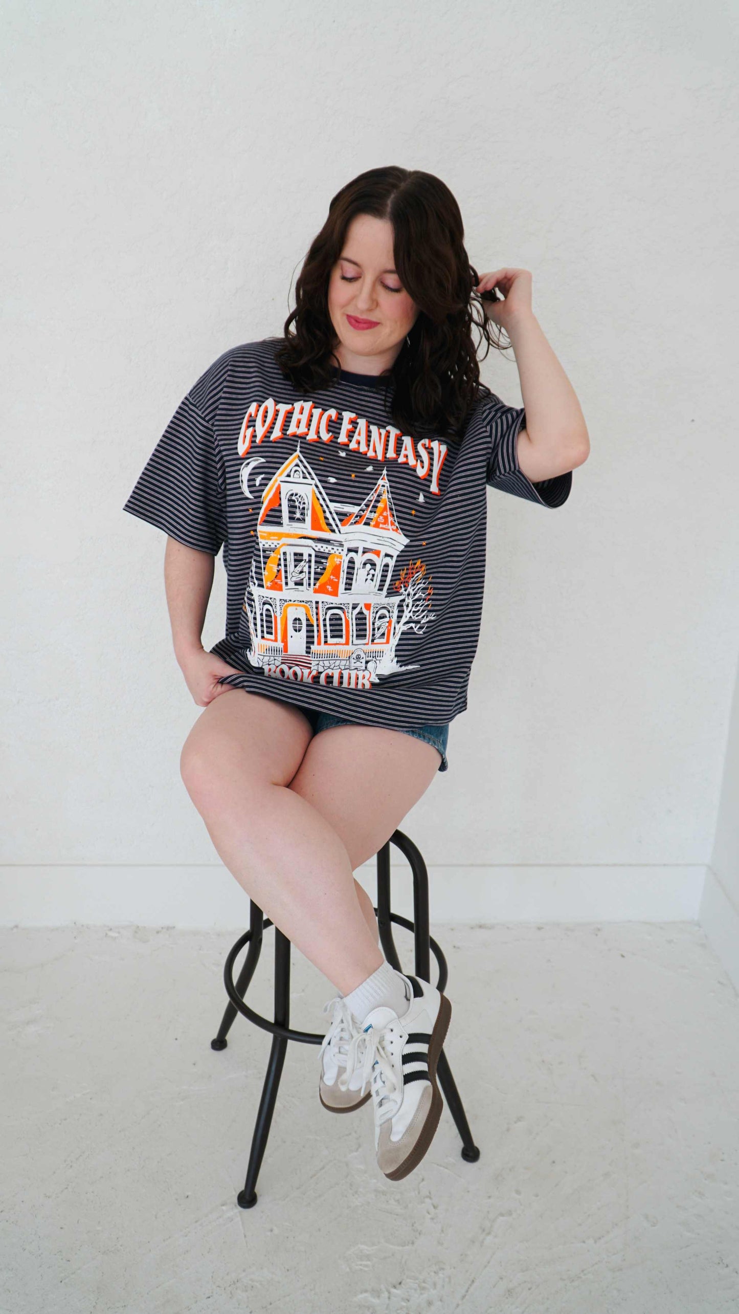Woman wearing a 'Gothic Fantasy' t-shirt sitting on a stool against a white background. Striped oversized boxy Gothic Fantasy Tee / t-shirt. For book lovers of the romantasy genre.