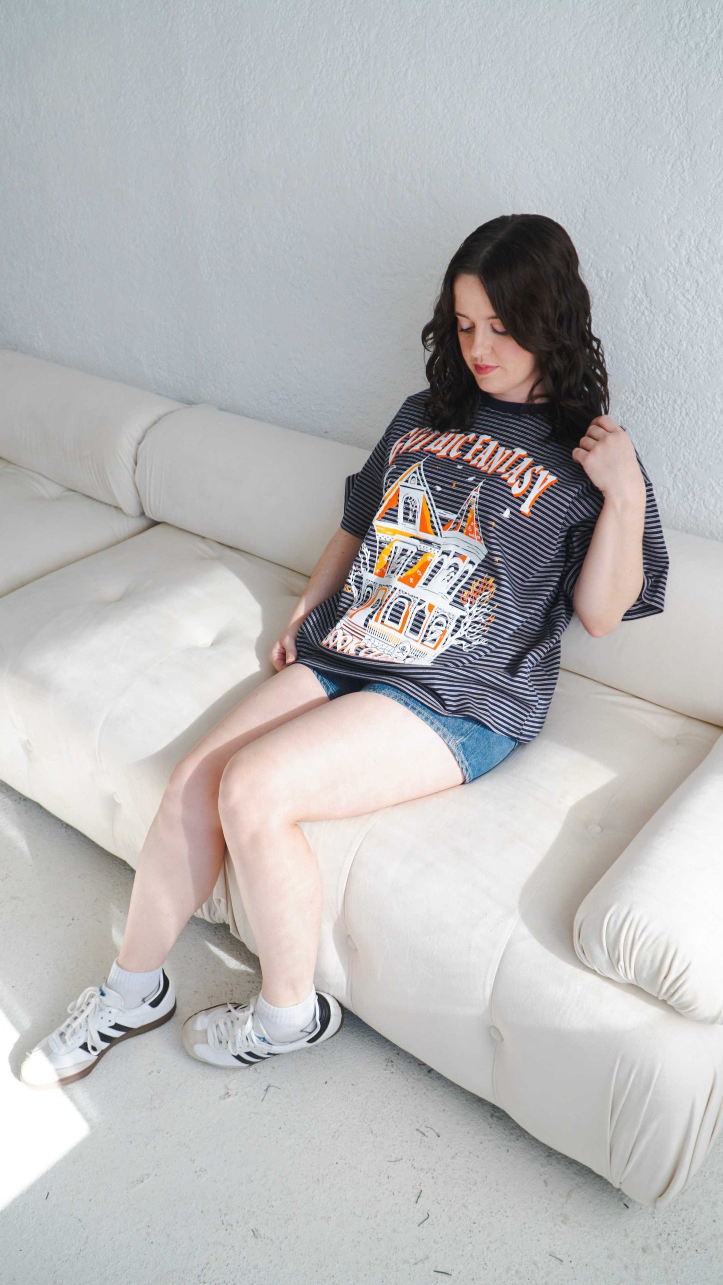 Woman sitting on a white couch wearing a graphic t-shirt and denim shorts. Striped oversized boxy Gothic Fantasy Tee / t-shirt. For book lovers of the romantasy genre.