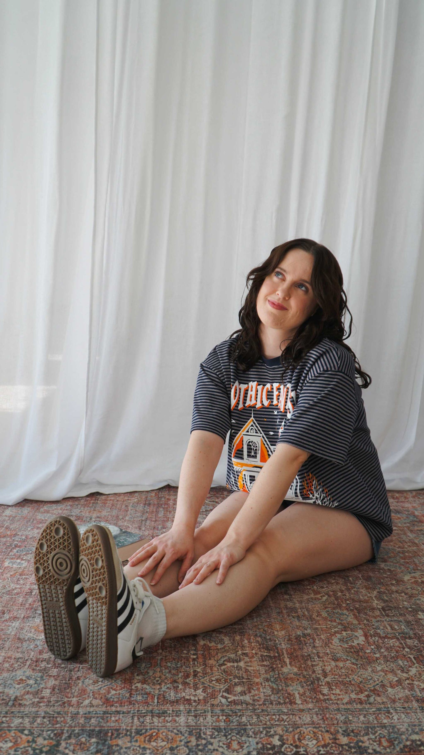 Woman sitting on a patterned rug with white curtains in the background. Striped oversized boxy Gothic Fantasy Tee / t-shirt. For book lovers of the romantasy genre.
