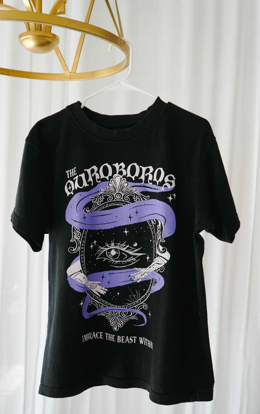 Black t-shirt with graphic design hanging on a white curtain background. A black graphic tee with 'The Ourororos' design featuring an eye, mirror, and purple mist. Inspired by the A Court of Thorns of Roses book series by Sarah J. Maas.