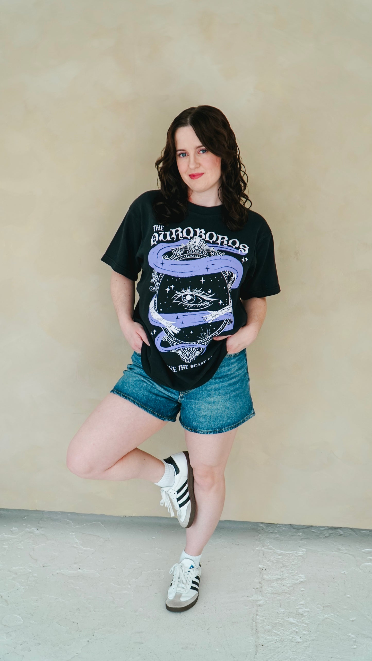Person wearing a black graphic t-shirt and denim shorts against a beige wall. She is wearing a black graphic tee with 'The Ourororos' design featuring an eye, mirror, and purple mist. Inspired by the A Court of Thorns of Roses book series by Sarah J. Maas.