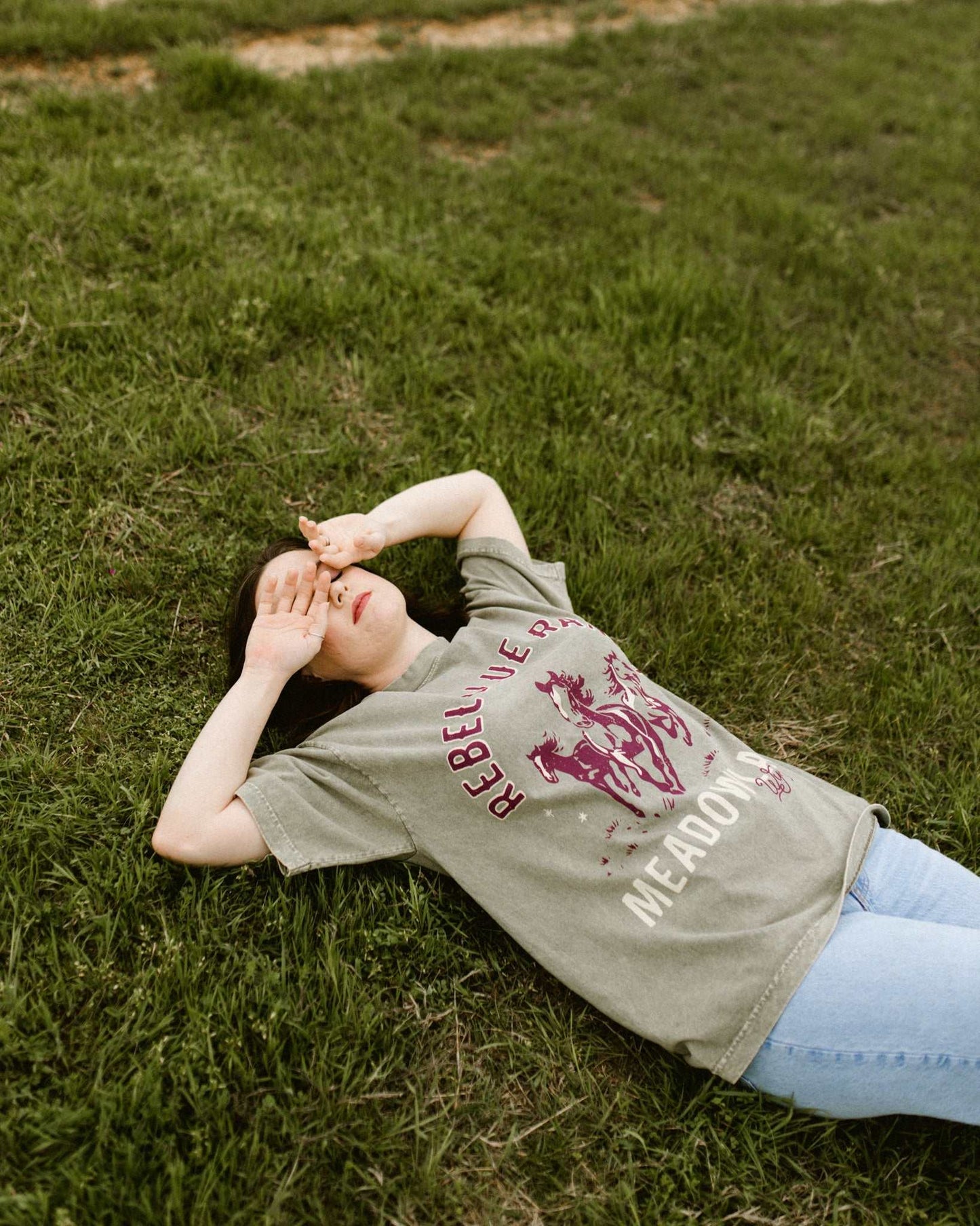 Rebel Blue Ranch Mustangs Tee. A woman wears a mineral wash olive green boxy oversized Rebel Blue Ranch tee with a horses design based on the cowboy romance book series by Lyla Sage that includes Done and Dusted, Swift and Saddled, Lost and Lassoed, and Wild and Wrangled. Meadowlark bookish merch. Lyla Sage merch.