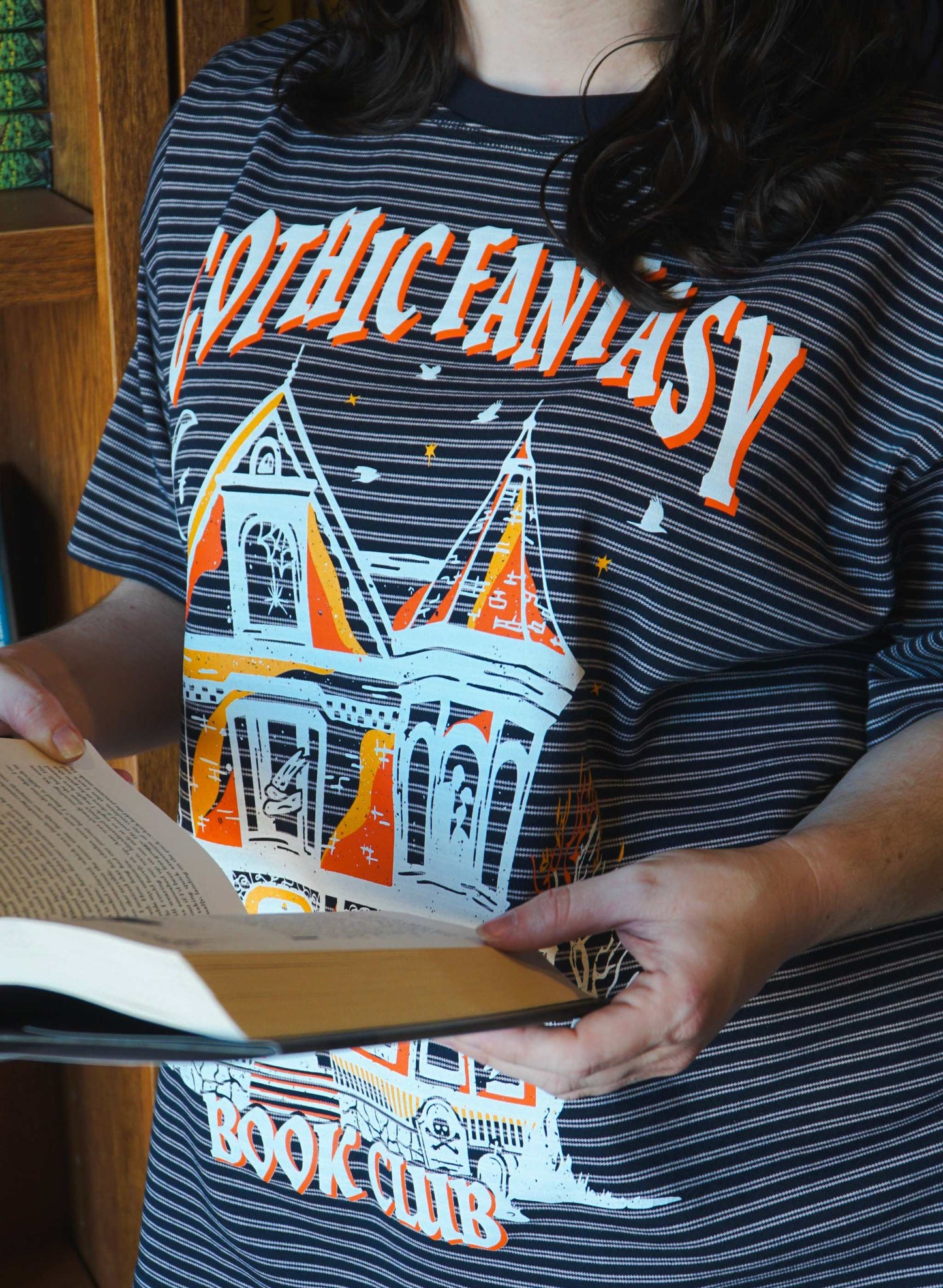 Person wearing a striped shirt with 'Gothic Fantasy Book Club' design, holding an open book. Striped oversized boxy Gothic Fantasy Tee / t-shirt. For book lovers of the romantasy genre.