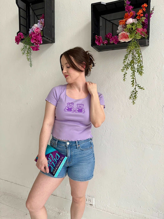 The Book Bears Baby Tee features a design with two teddy bears each holding a book. The t-shirt is a light purple color with a deep purple bookish design and lettuce hem trim.