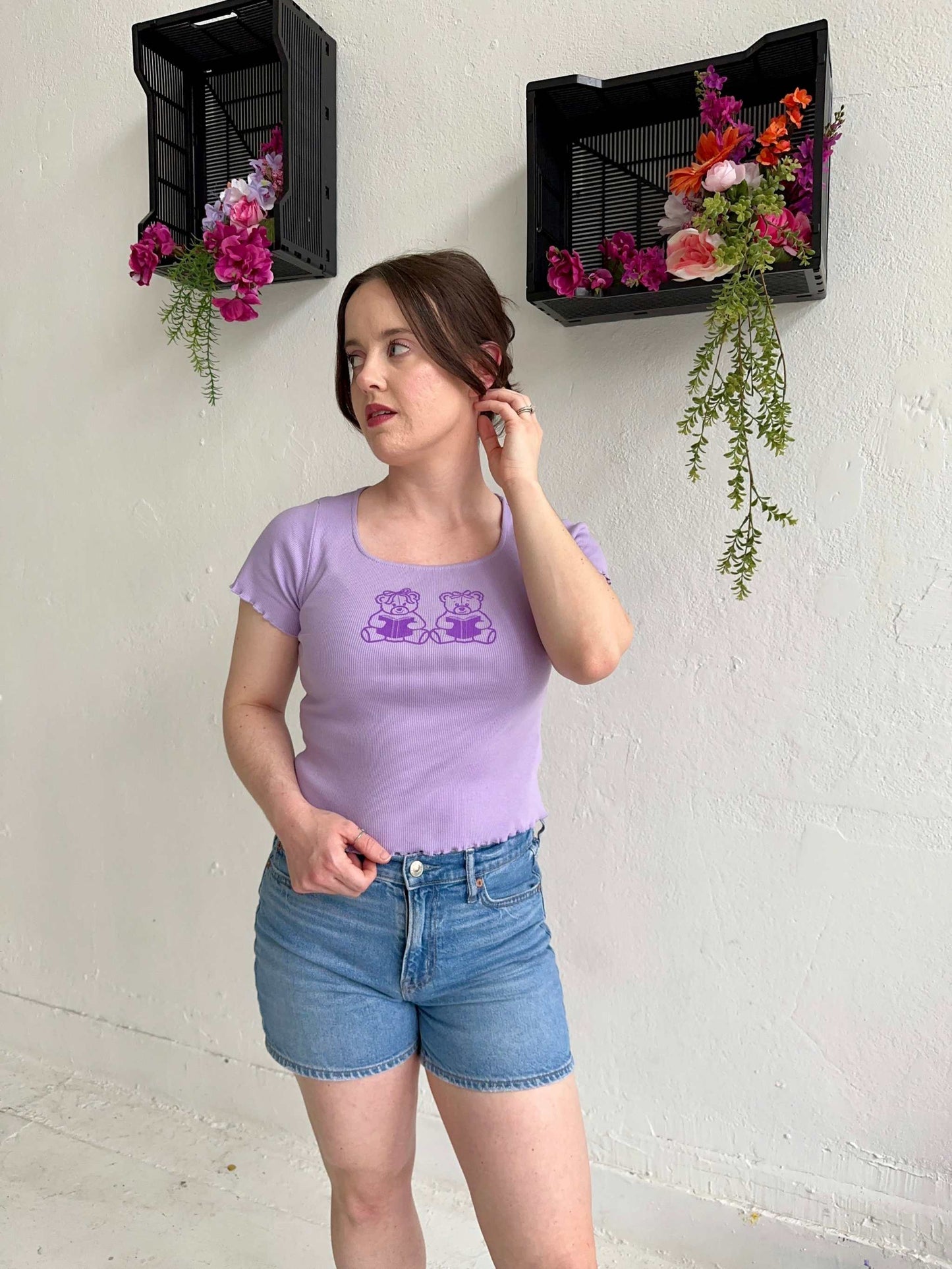 The Book Bears Baby Tee features a design with two teddy bears each holding a book. The t-shirt is a light purple color with a deep purple bookish design and lettuce hem trim.