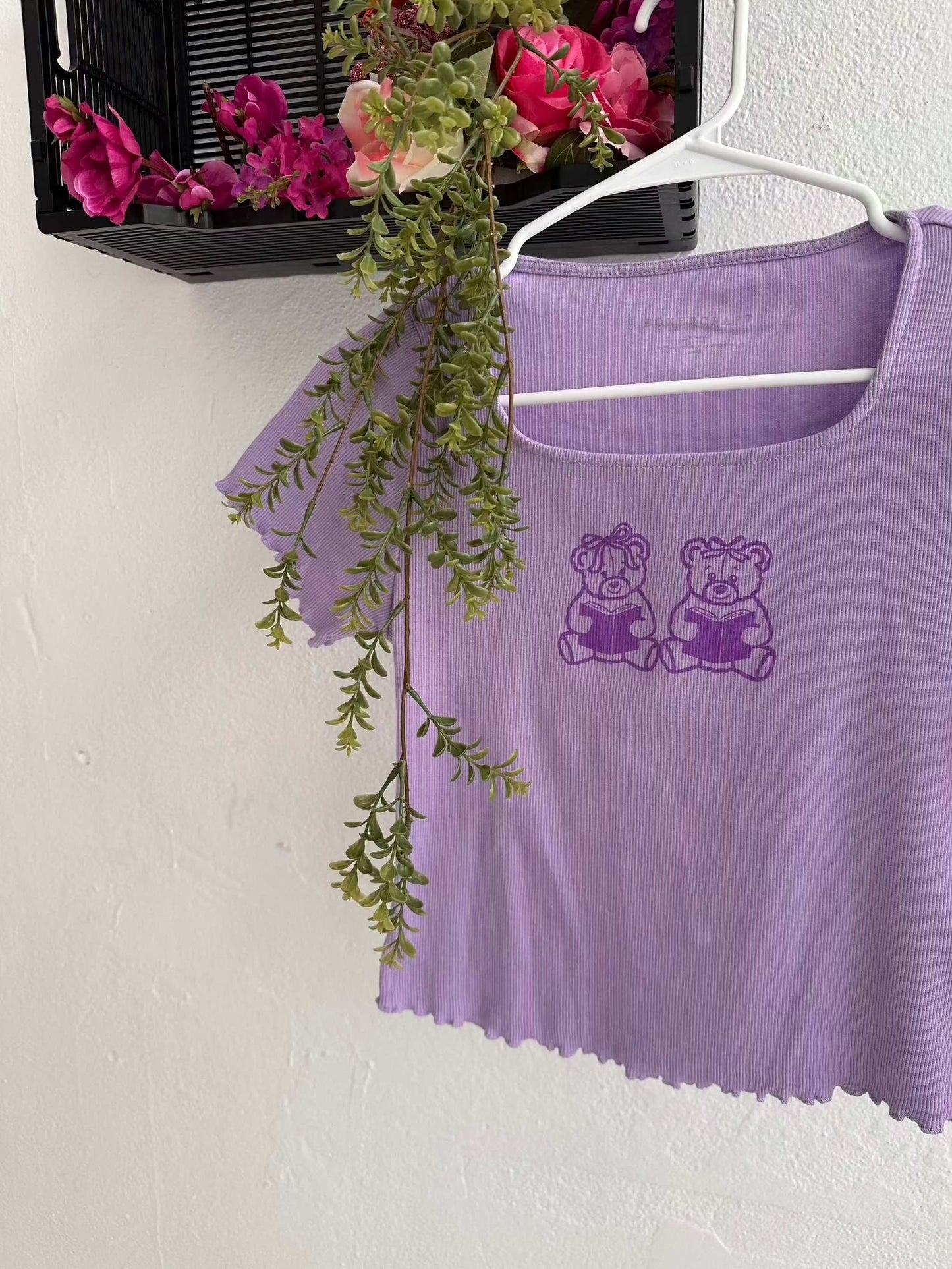 Purple baby tee with Book Bears design, lettuce trim hem, hanging with floral decor.