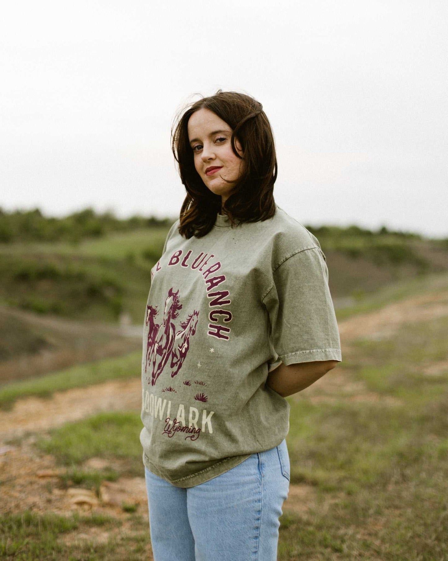Rebel Blue Ranch Mustangs Tee. A woman wears a mineral wash olive green boxy oversized Rebel Blue Ranch tee with a horses design based on the cowboy romance book series by Lyla Sage that includes Done and Dusted, Swift and Saddled, Lost and Lassoed, and Wild and Wrangled. Meadowlark bookish merch. Lyla Sage merch.