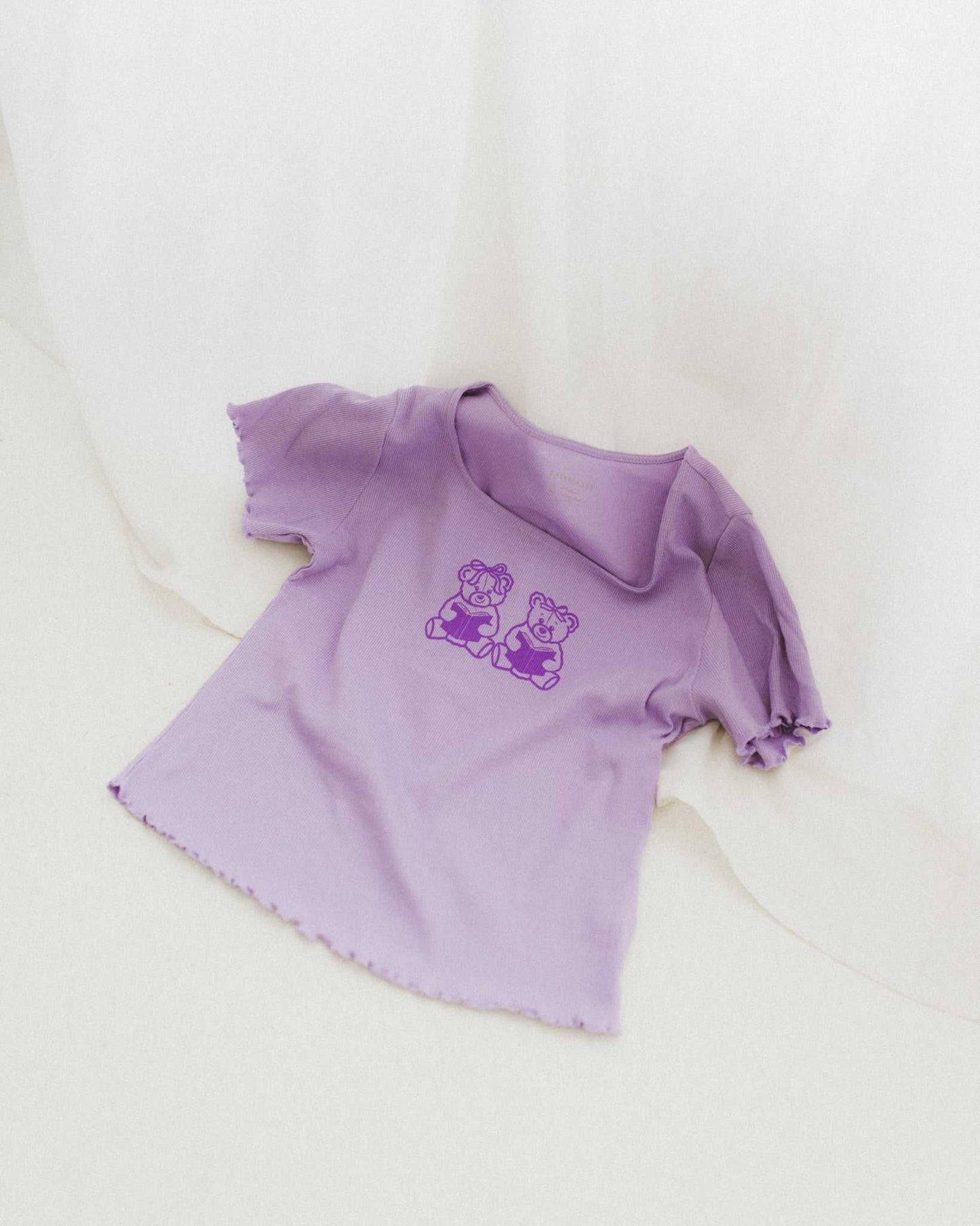 The Book Bears Baby Tee features a design with two teddy bears each holding a book. The t-shirt is a light purple color with a deep purple bookish design and lettuce hem trim.