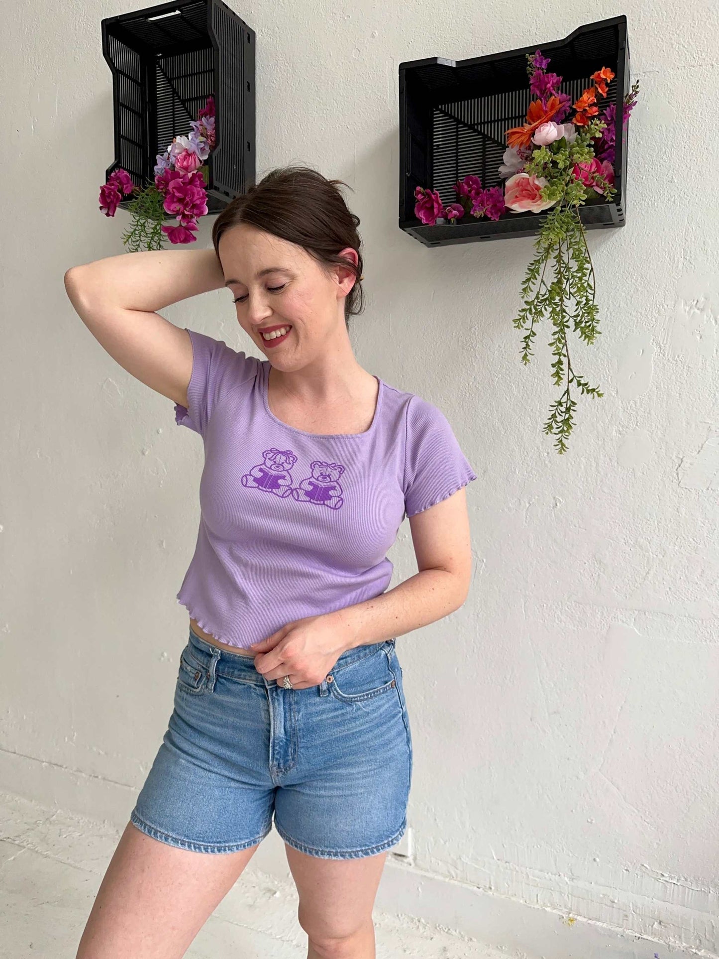 The Book Bears Baby Tee features a design with two teddy bears each holding a book. The t-shirt is a light purple color with a deep purple bookish design and lettuce hem trim.
