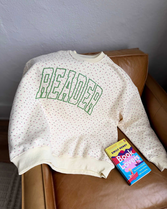 The Reader Floral Crewneck from Next Chapter features a floral patterned fabric with a green puff print 'Reader' design on the front. Next Chapter offers high quality bookish apparel.