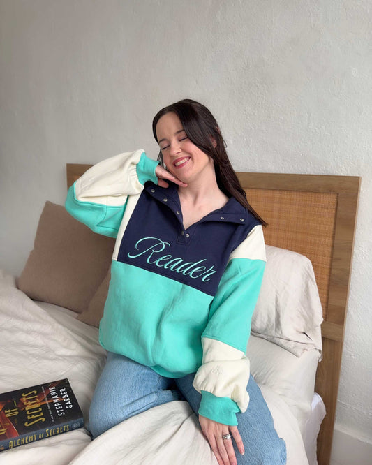 The Reader Colorblock Pullover from Next Chapter is oversized, with a quarter button-up style. This variant is navy, teal, and ivory and has the word 'Reader' embroidered across the front. Next Chapter offers high quality bookish merch and apparel.