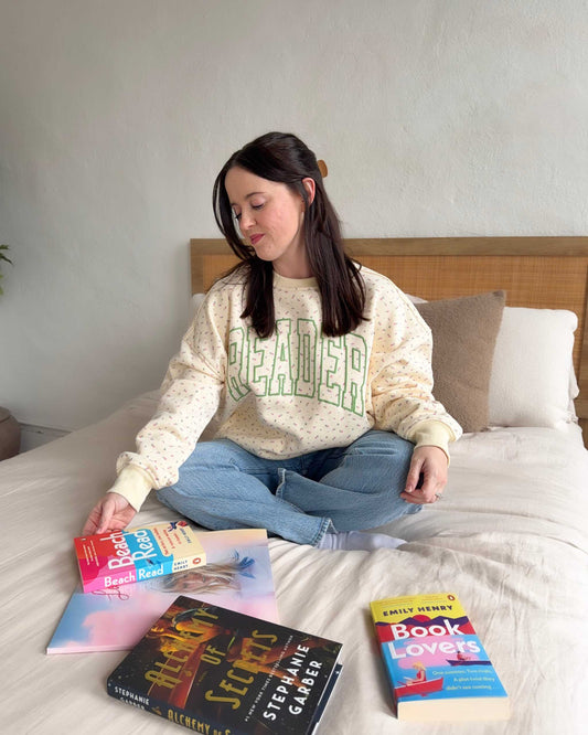 The Reader Floral Crewneck from Next Chapter has a pale yellow fabric with a subtle floral pattern. Across the sweatshirt is the word 'READER' screen printed in green. Next Chapter offers high quality bookish merch and apparel.