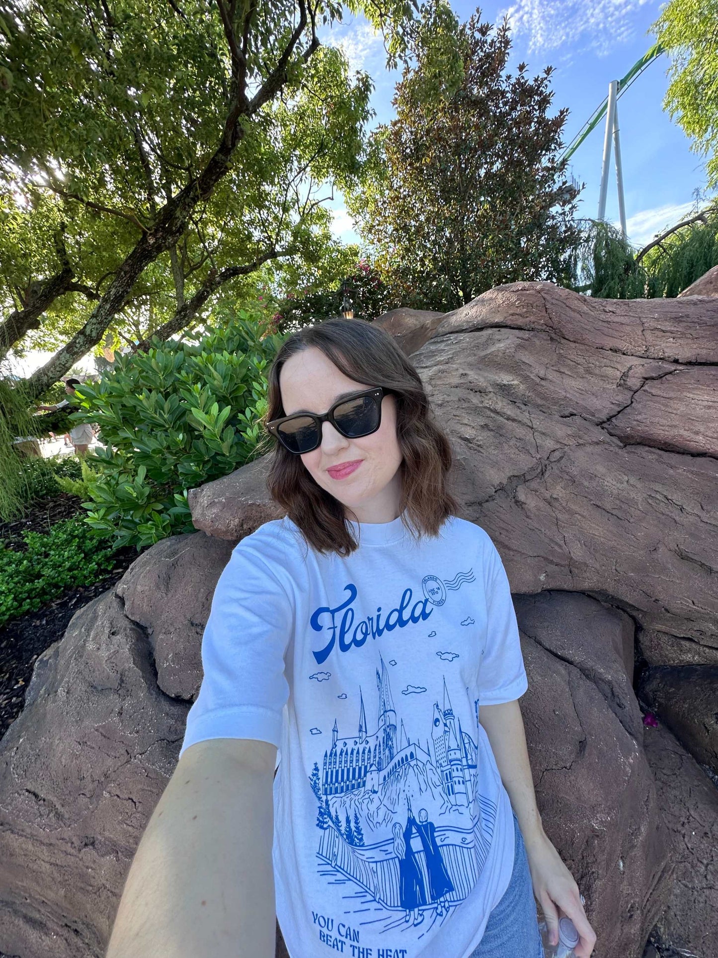 Florida Wizarding Parks Tee. A woman wears a t-shirt that depicts the Wizarding World of Harry Potter theme park. The t-shirt also references Taylor Swift's song "Florida!!!" as well as the opening date of the park.