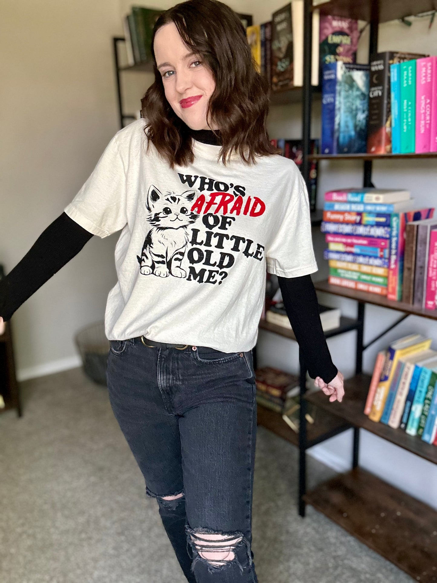 A woman is standing in front of a bookshelf wearing a t-shirt with a Taylor Swift lyric. The shirt has a kitten on it with the quote "who's afraid of little old me?" The shirt is ivory with a black and red design.