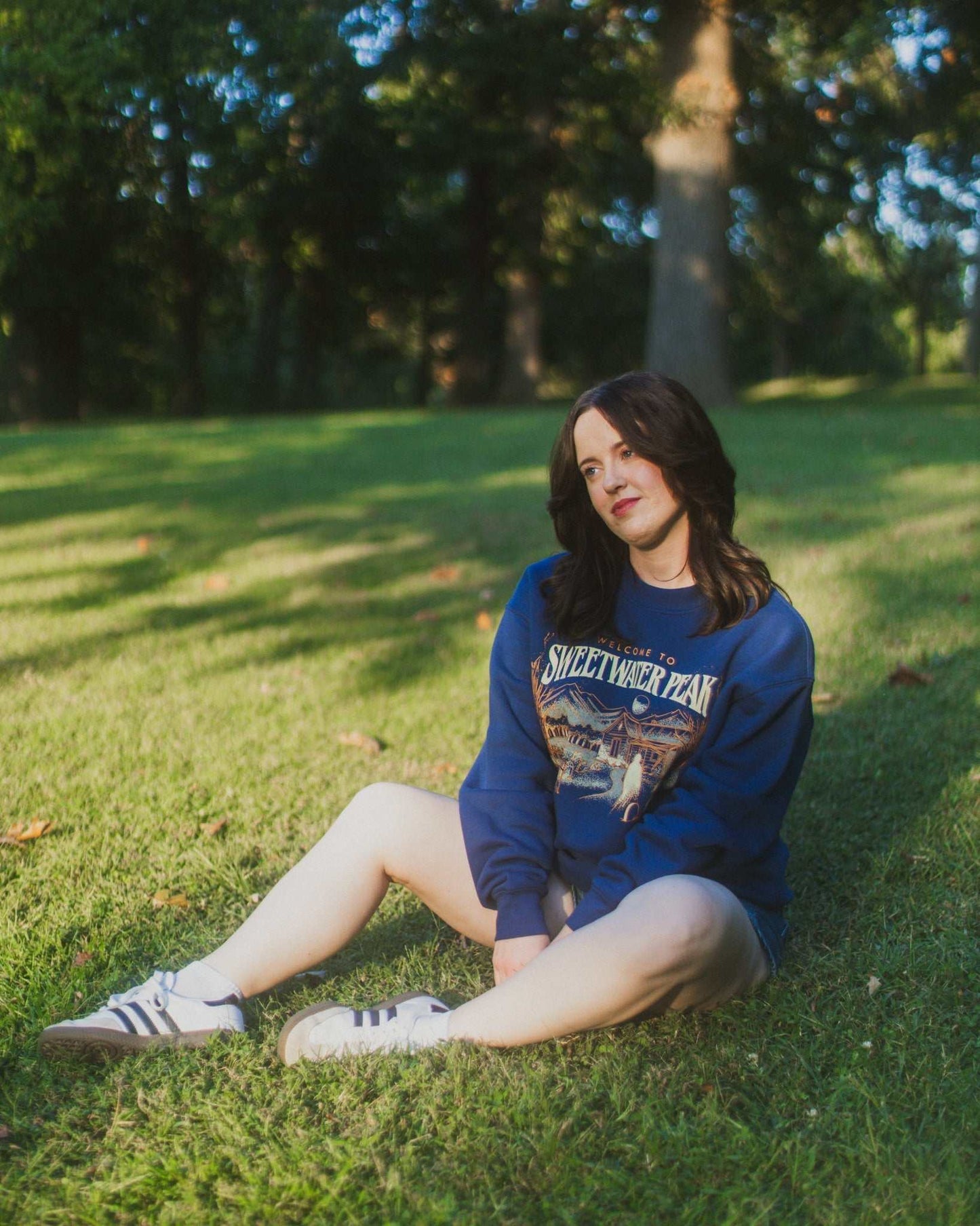 Person sitting on grass wearing a blue sweatshirt with a graphic design. The Sweetwater Peak crewneck design is inspired by the Lyla Sage book, Soul Searching, which is from her new Sweetwater Peak book series. Merch for book lovers. Officially licensed by Lyla Sage.
