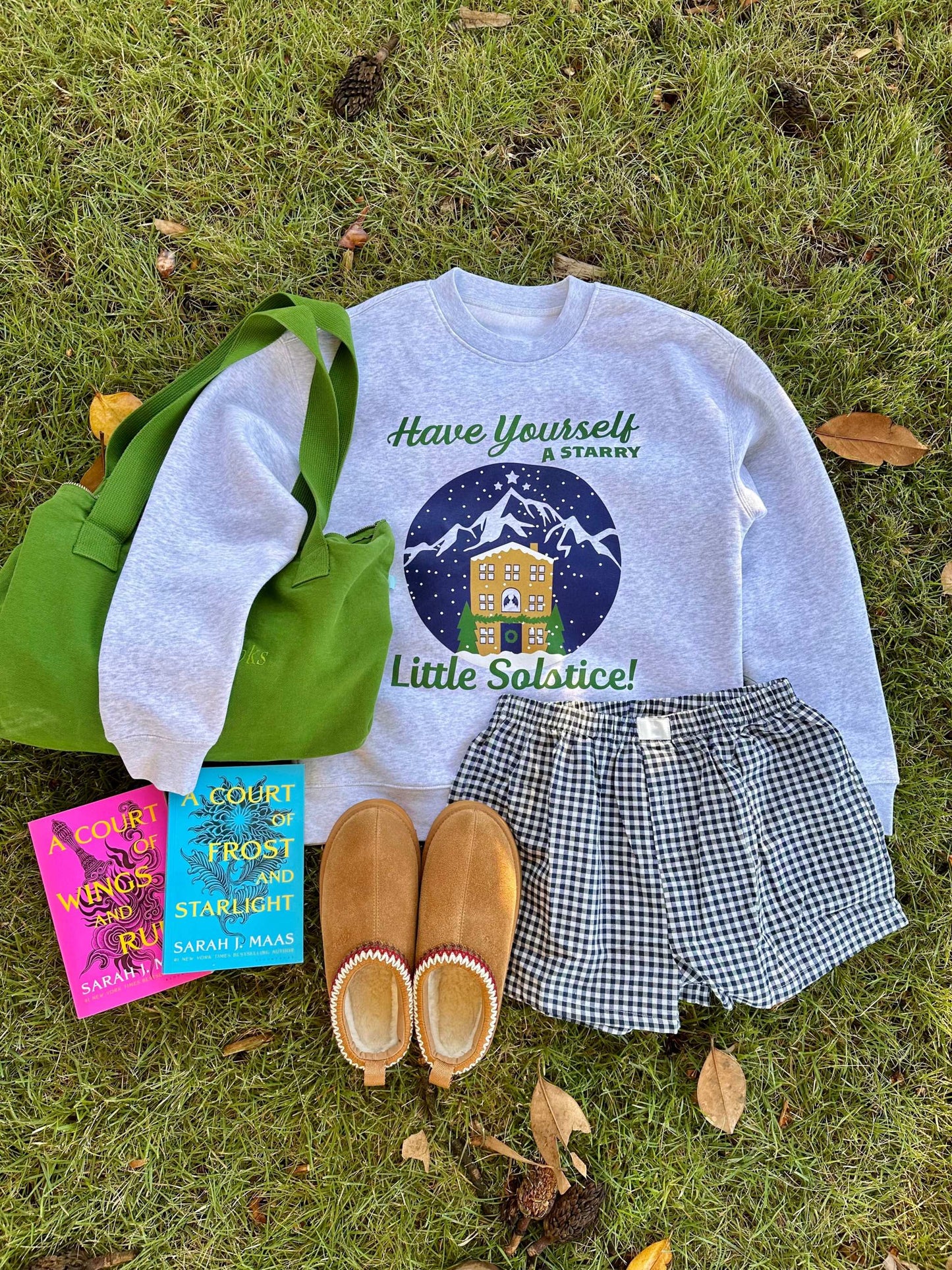 Gray Starry Solstice Crewneck sweatshirt with holiday design, paired with green tote, checkered shorts, slippers, and two Sarah J. Maas books on grass