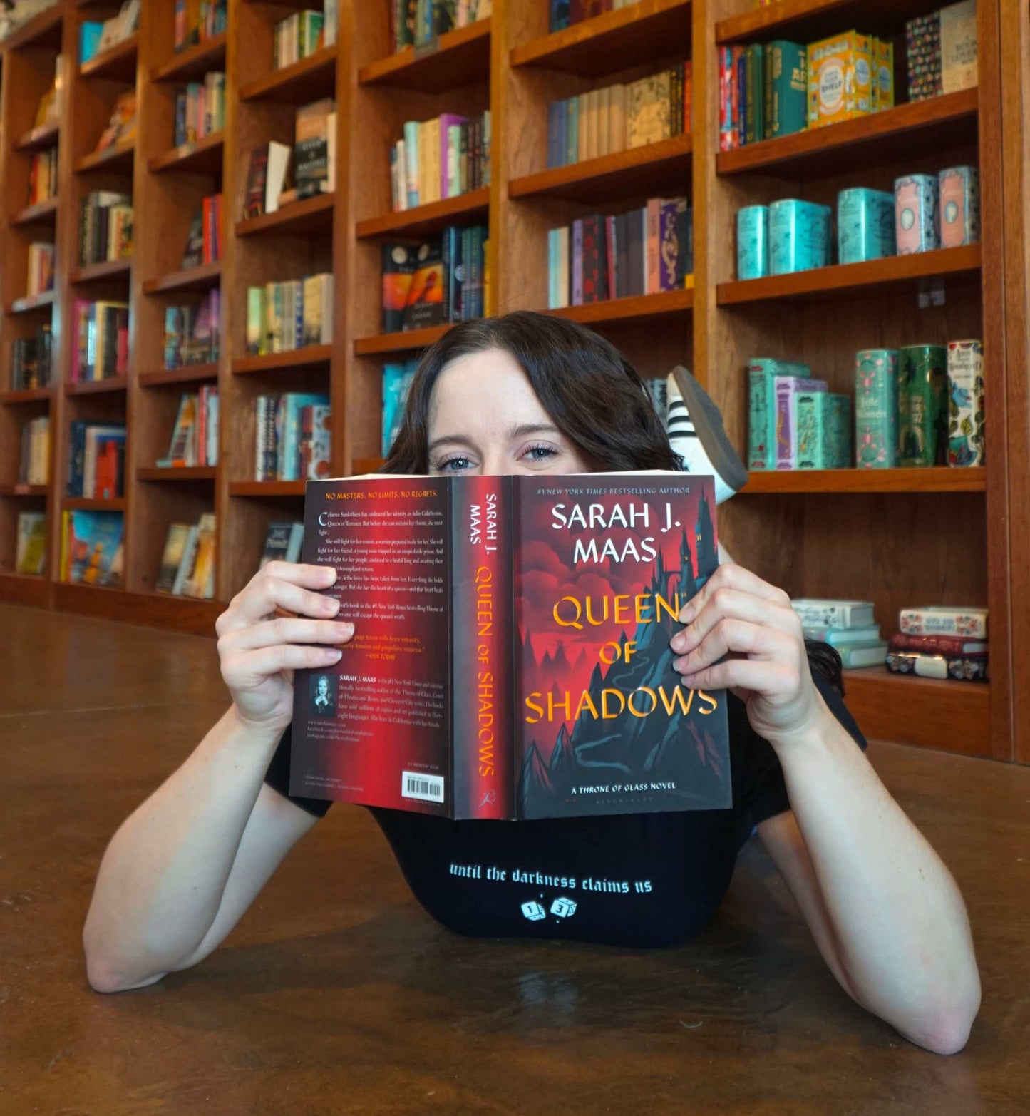 Person holding a book titled 'Queen of Shadows' by Sarah J. Maas in a library setting. The Thirteen Tee from Throne of Glass by Sarah J. Maas. "Until the darkness claims us" Ironteeth Witches design.