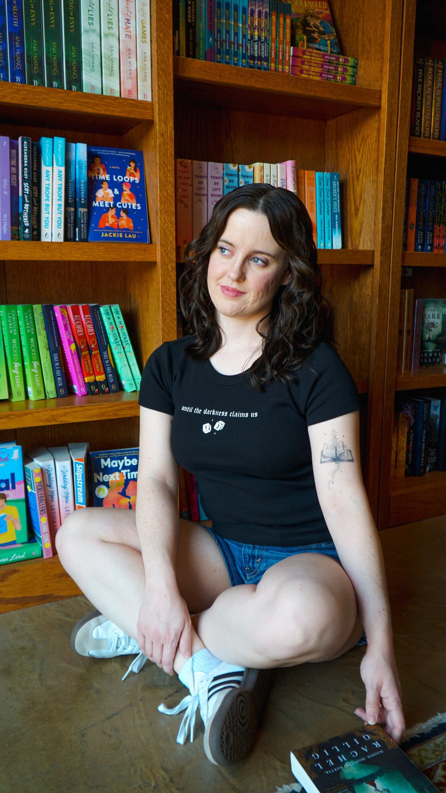 Woman sitting on the floor in front of a bookshelf filled with books. The Thirteen Tee from Throne of Glass by Sarah J. Maas. "Until the darkness claims us" Ironteeth Witches design.