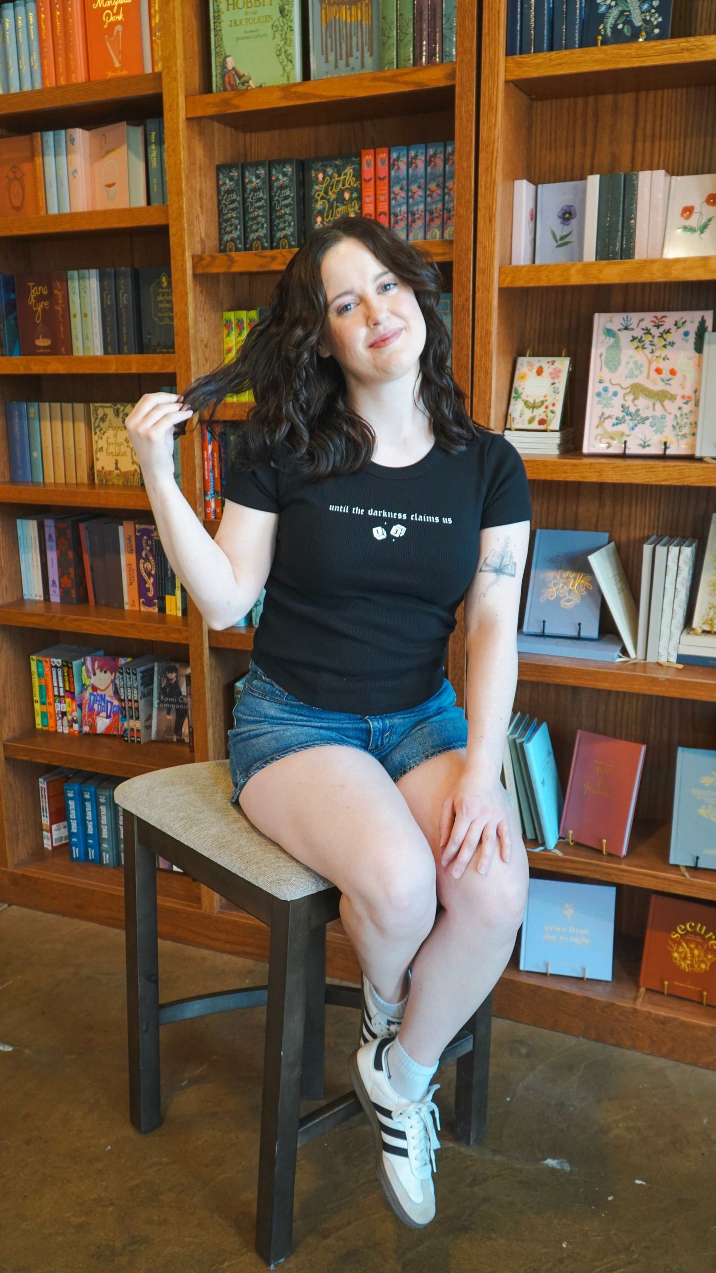Woman sitting on a chair in front of bookshelves with books. The Thirteen Tee from Throne of Glass by Sarah J. Maas. "Until the darkness claims us" Ironteeth Witches design.
