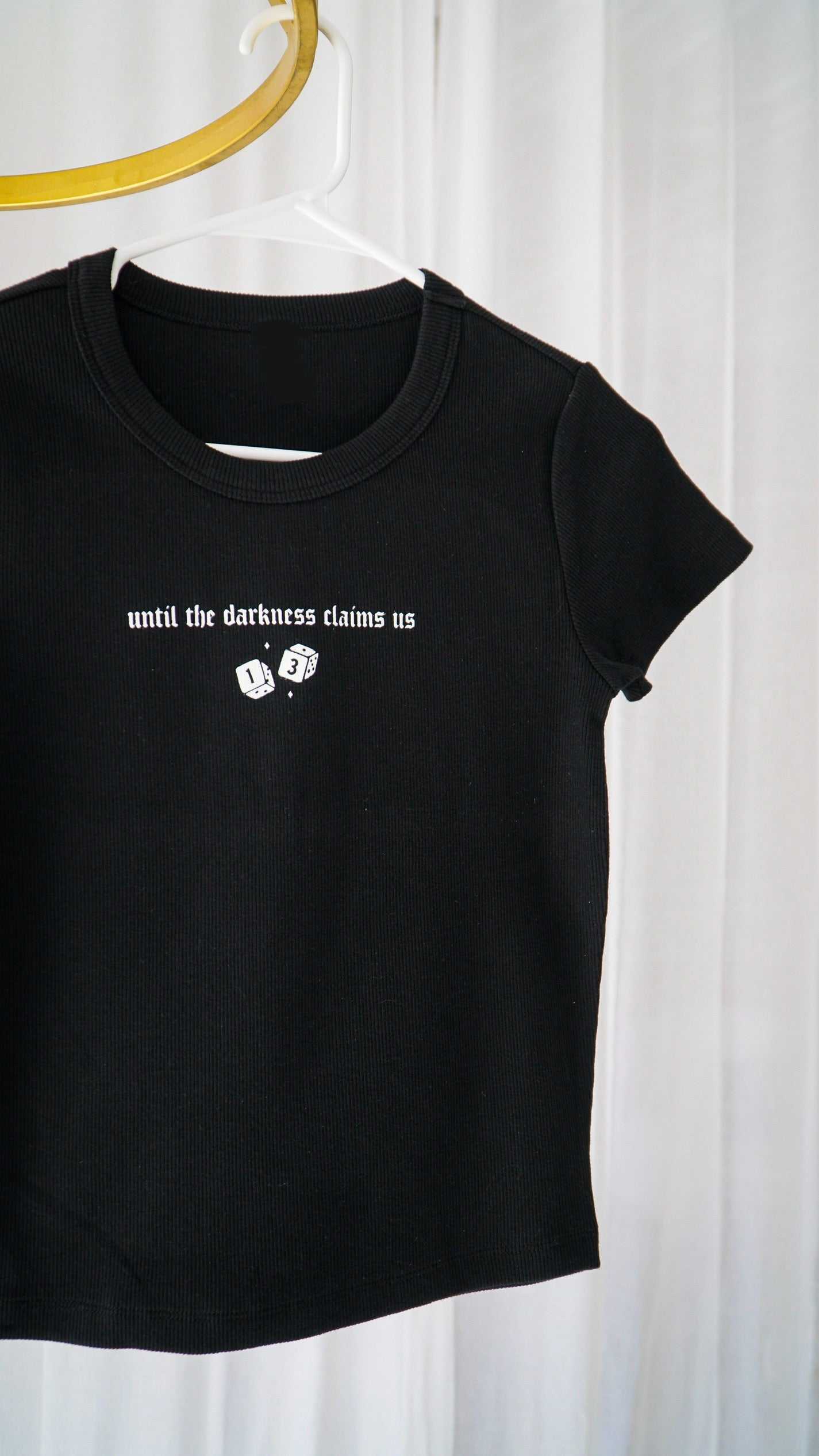 Black t-shirt with white text and graphics on a hanger against a white curtain background. The Thirteen Tee from Throne of Glass by Sarah J. Maas. "Until the darkness claims us" Ironteeth Witches design.