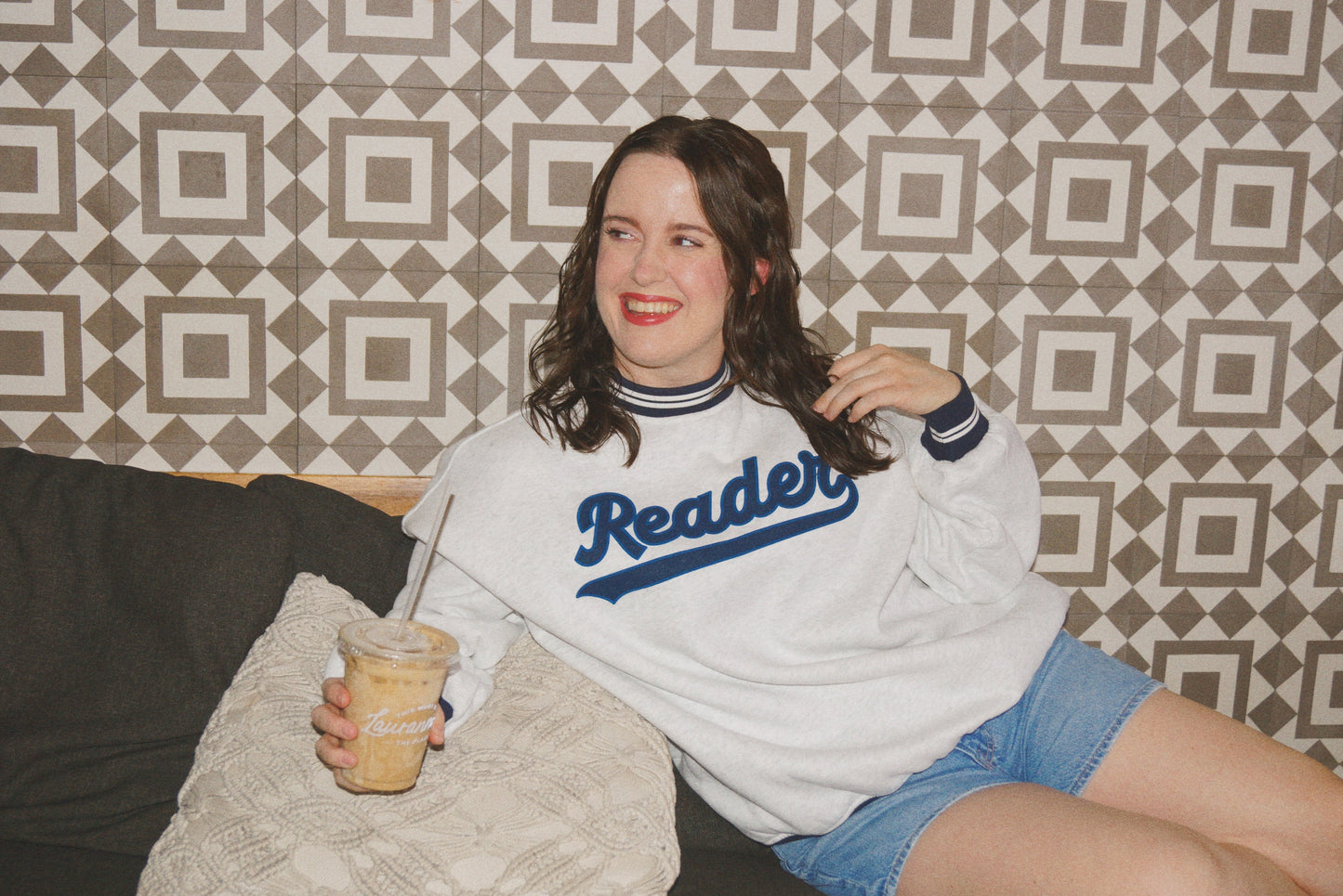 Woman wearing ash gray Reader Crewneck with navy striped collar and cuffs, holding iced coffee and sitting on patterned couch