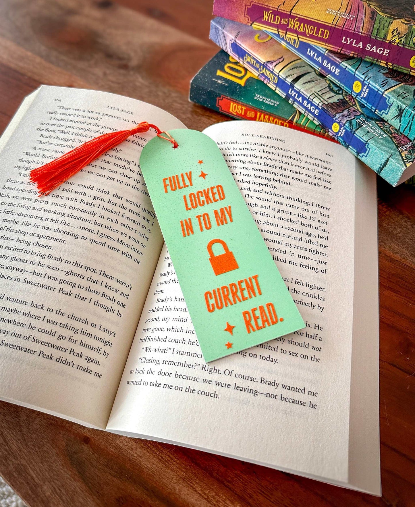 Open book with a mint green bookmark reading 'Fully Locked In To My Current Read' on a wooden surface. Created for the bookish swifties to celebrate The Life of a Showgirl.