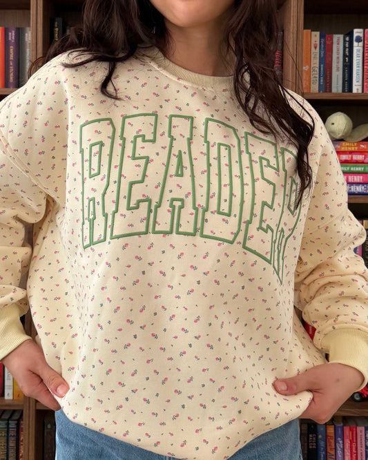 The Reader Floral Crewneck from Next Chapter has a pale yellow fabric with a subtle floral pattern. Across the sweatshirt is the word 'READER' screen printed in green. Next Chapter offers high quality bookish merch and apparel.