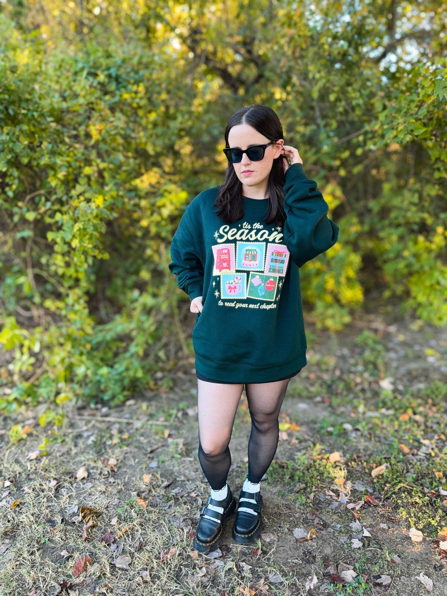 Woman wearing a festive Next Chapter Holiday Crewneck Sweatshirt with bookish Christmas-themed graphics, holding a phone, standing outdoors.