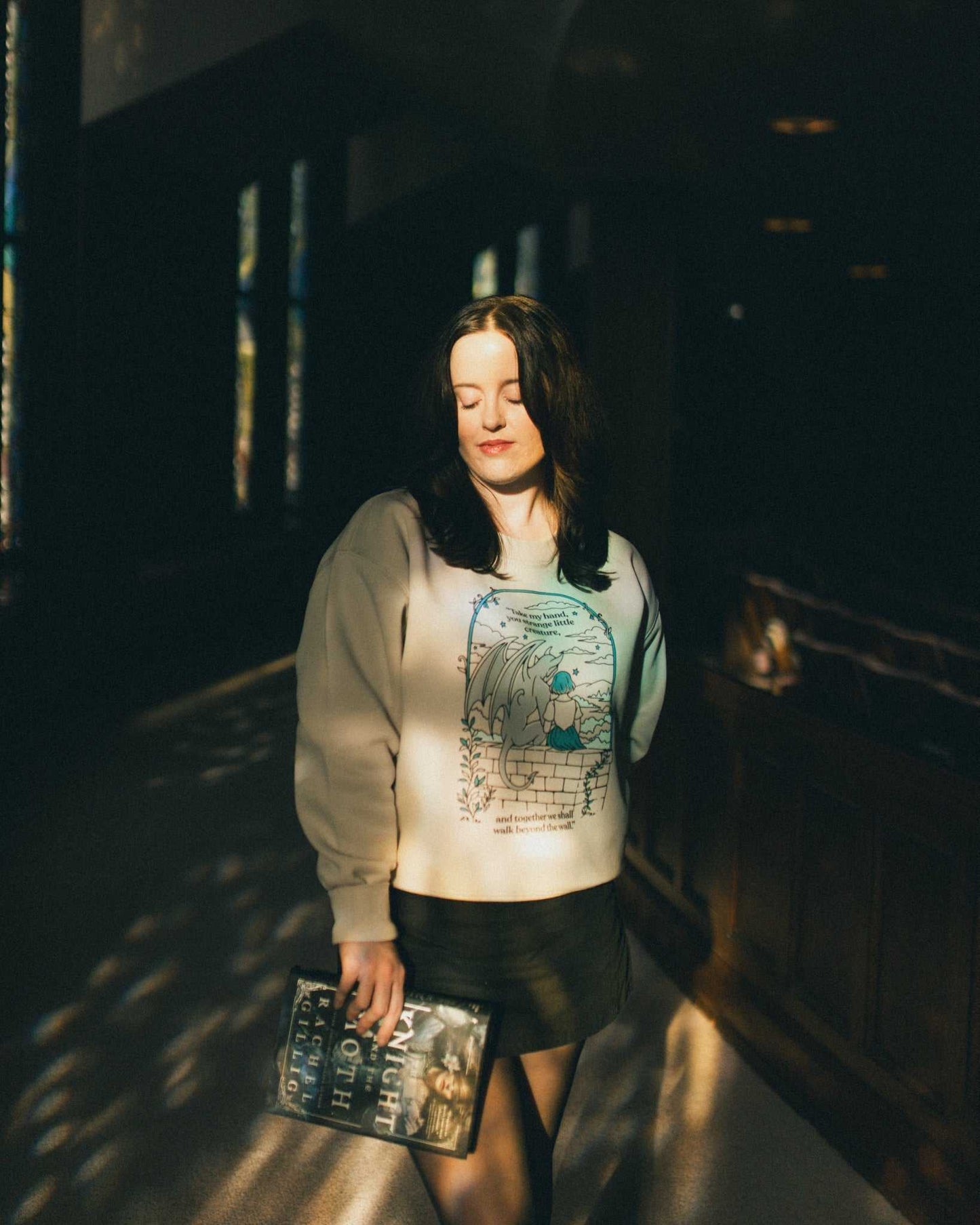 Woman wearing an ivory strange little creatures crewneck sweatshirt with a graphic design inspired by The Knight and the Moth by Rachel Gillig.