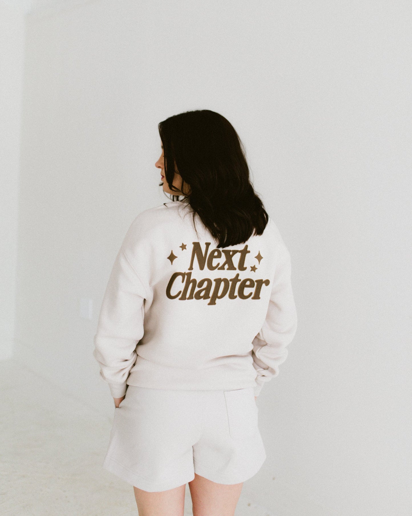 Ivory Next Chapter Crewneck sweatshirt with chocolate brown print worn by woman from back, relaxed fit with ribbed cuffs and crew neckline.