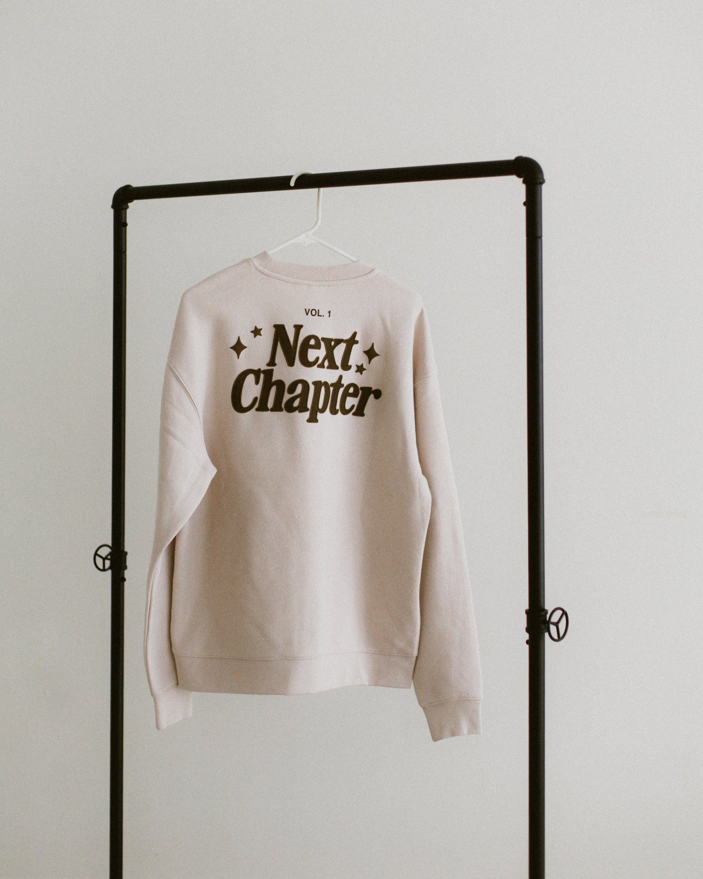 Ivory Next Chapter Crewneck sweatshirt with brown screen print and embroidery hanging on black clothing rack