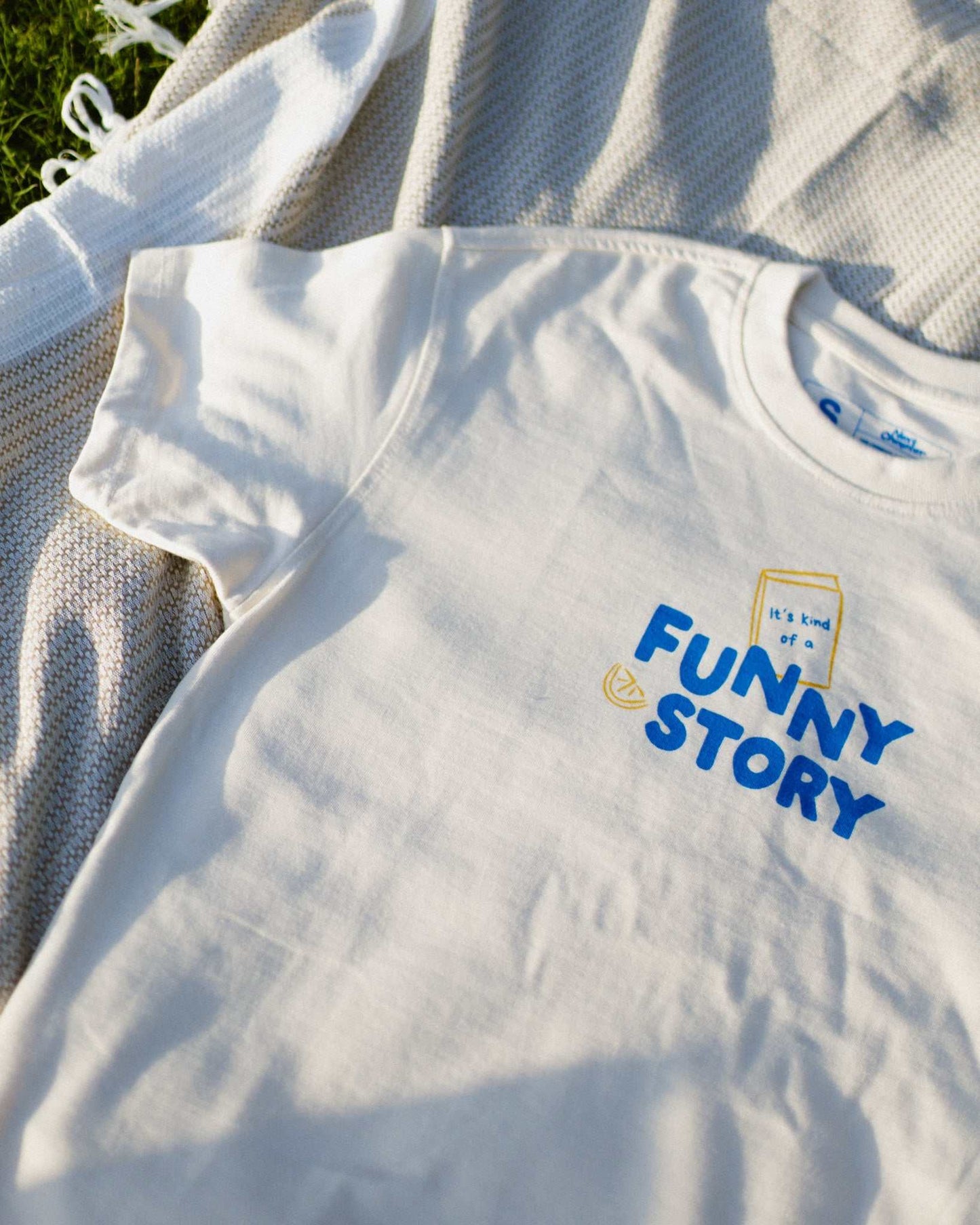 An oversized, boxy t-shirt, the Funny Story Tee is inspired by the book Funny Story by Emily Henry. The item is an ivory/cream color with a royal blue and gold design on the front and back.