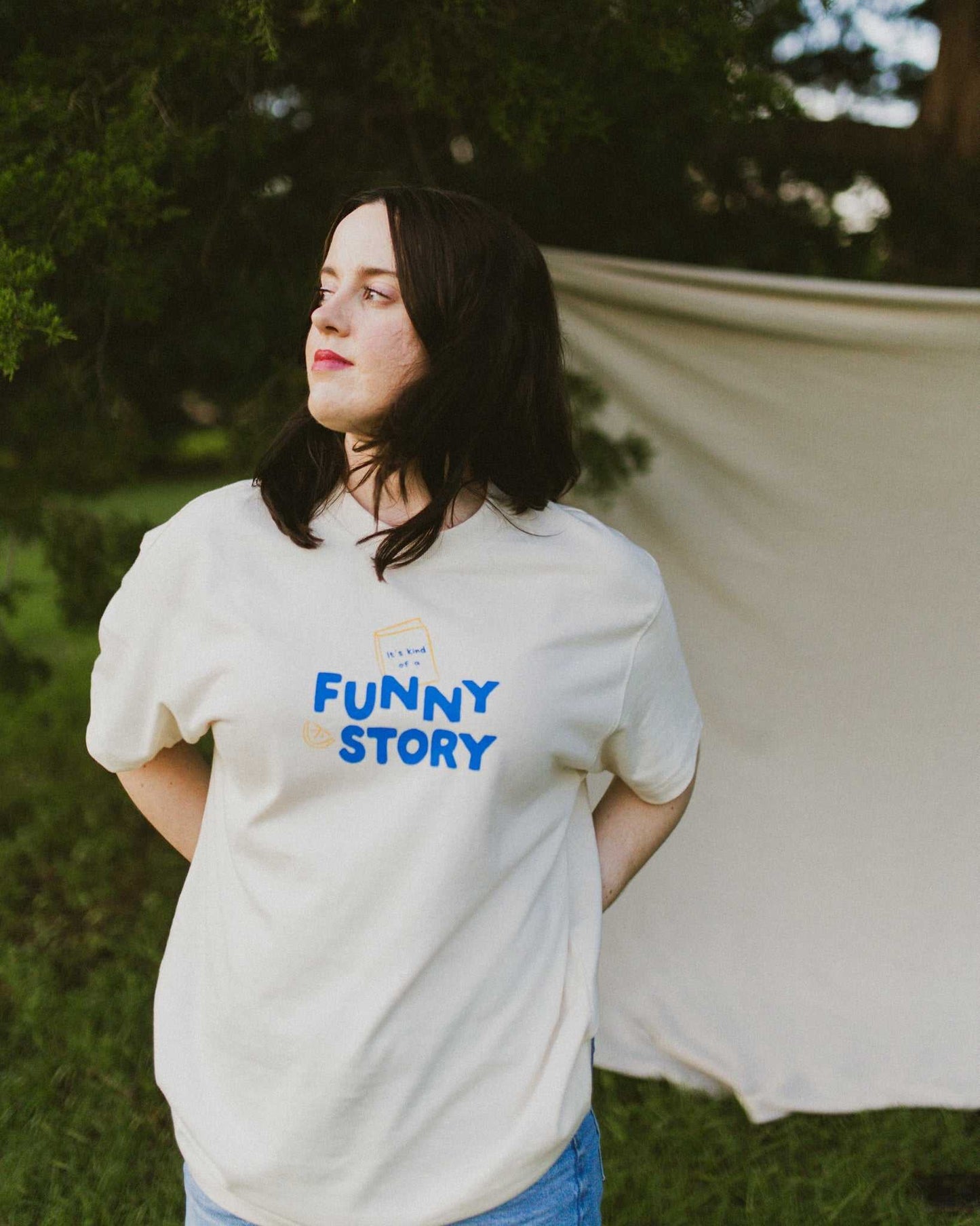 An oversized, boxy t-shirt, the Funny Story Tee is inspired by the book Funny Story by Emily Henry. The item is an ivory/cream color with a royal blue and gold design on the front and back.