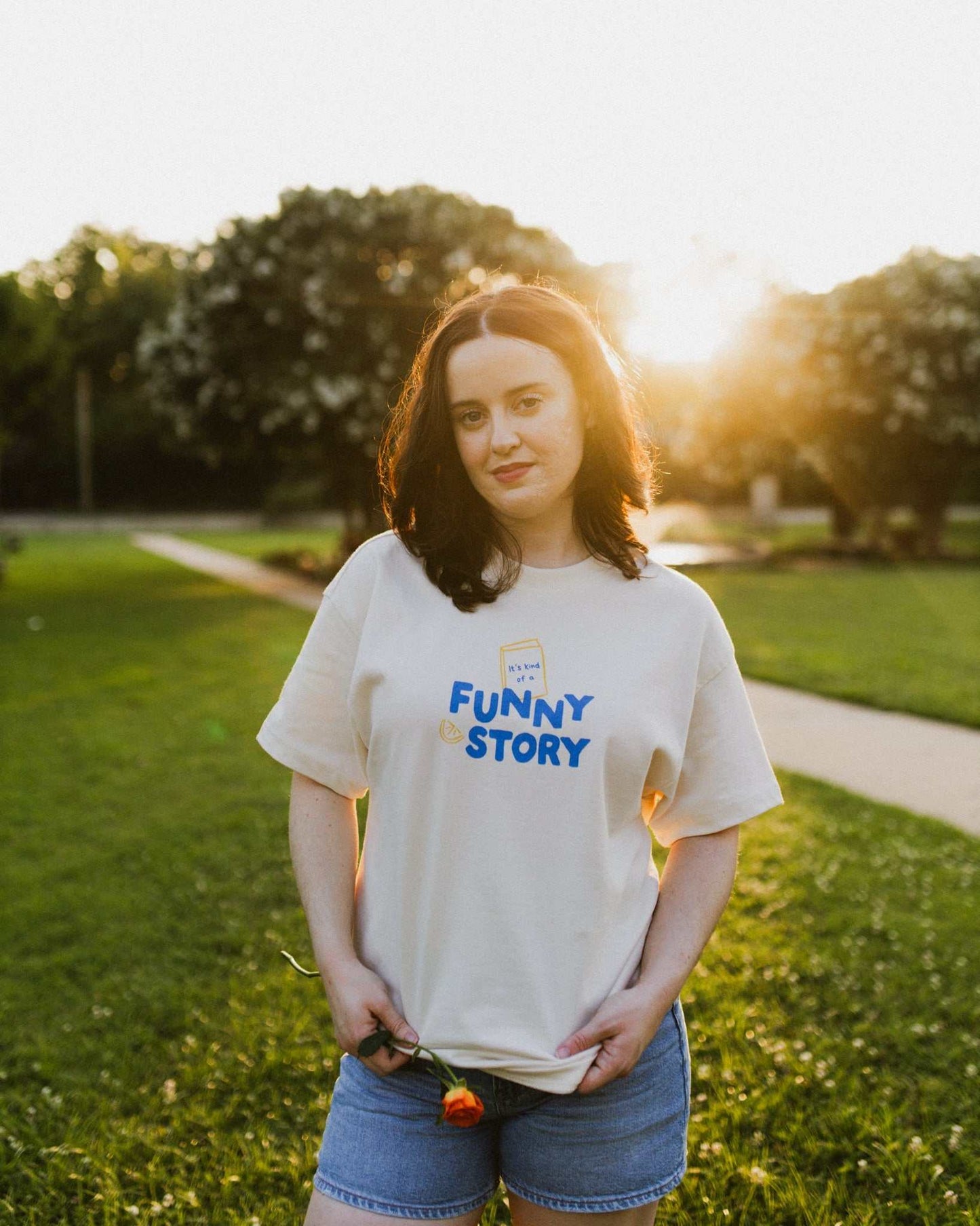 An oversized, boxy t-shirt, the Funny Story Tee is inspired by the book Funny Story by Emily Henry. The item is an ivory/cream color with a royal blue and gold design on the front and back.