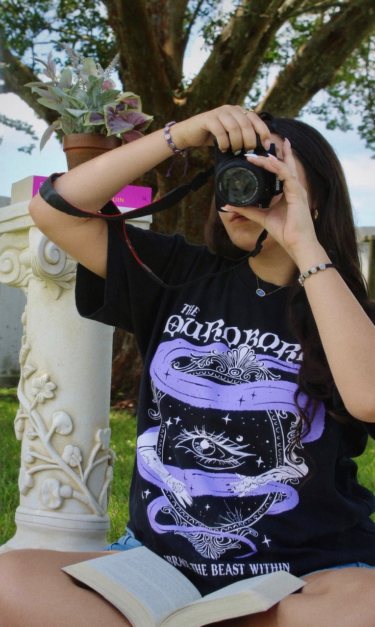 Person taking a photo with a camera, wearing a black t-shirt with a purple and white design. She is wearing a black graphic tee with 'The Ourororos' design featuring an eye, mirror, and purple mist. Inspired by the A Court of Thorns of Roses book series by Sarah J. Maas.