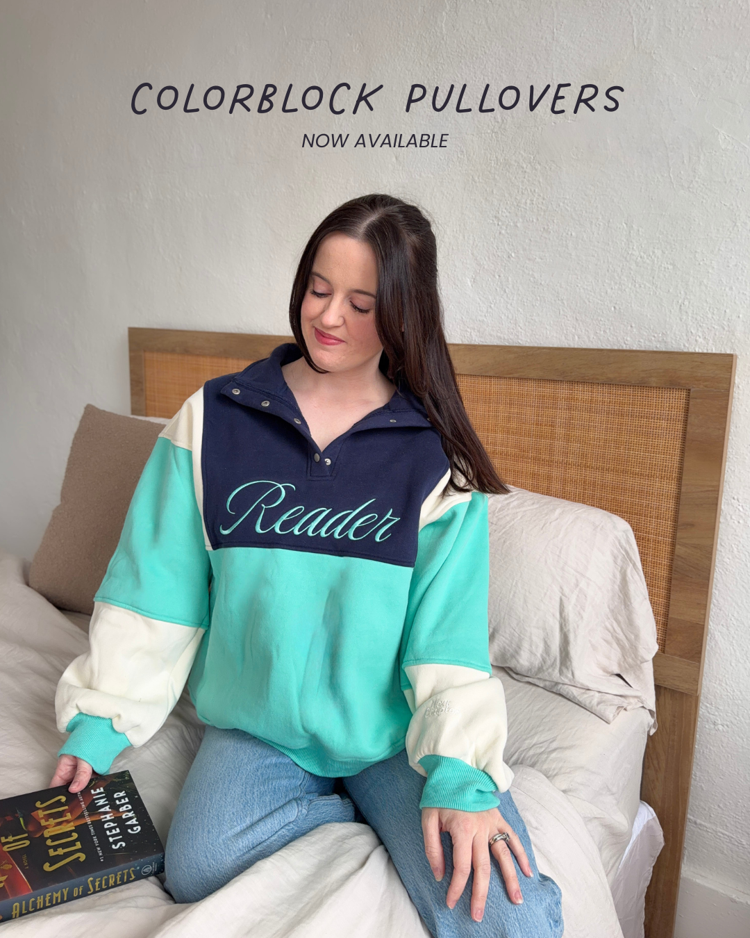 Woman wearing a colorblock pullover with 'Reader' text, sitting on a bed with text on the image that says 'now available'