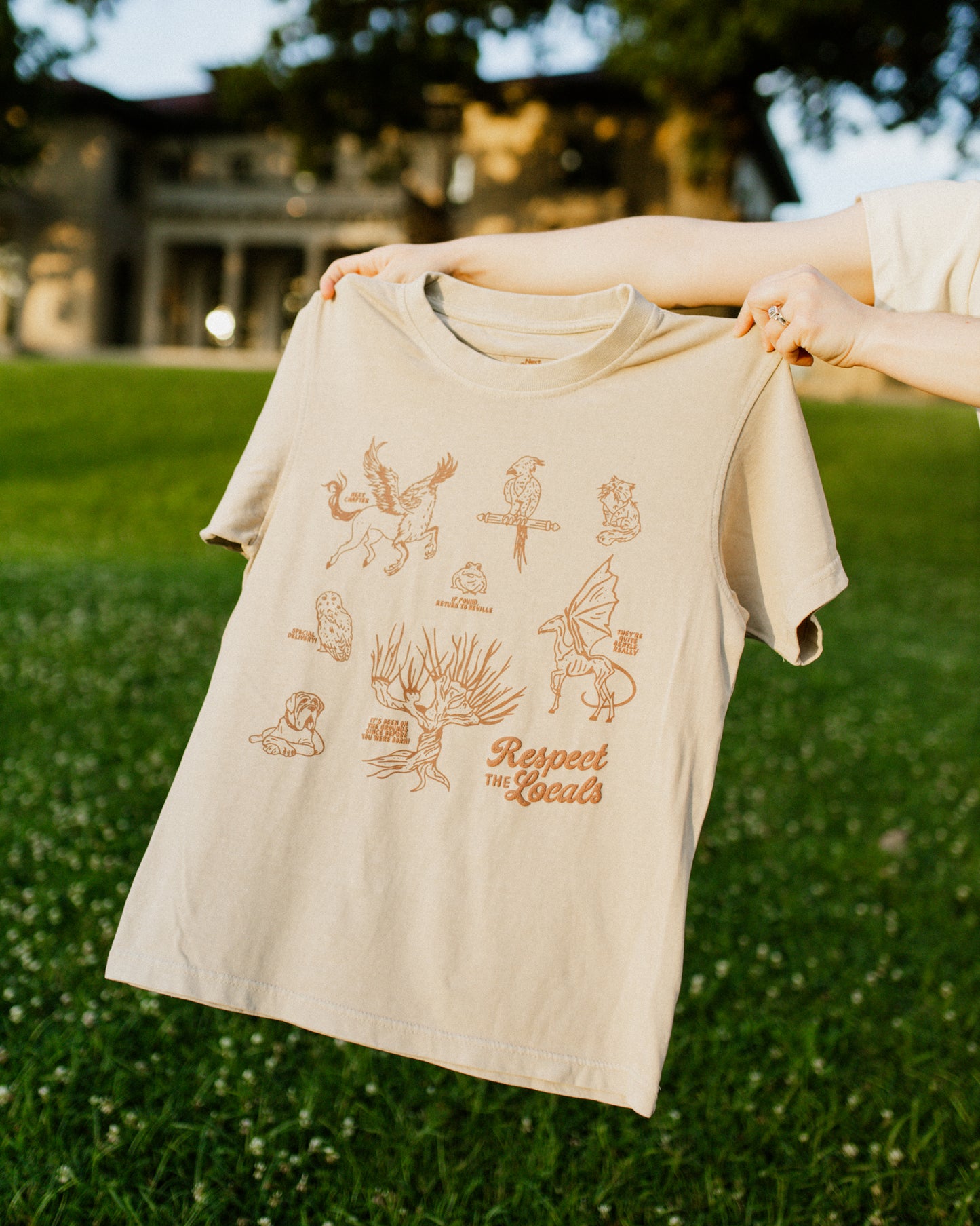 Beige t-shirt with dark brown graphic designs held by a person outdoors on grass. The design is inspired by the creatures in the Harry Potter universe.