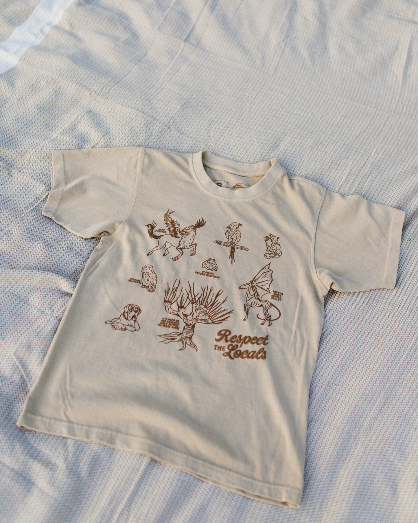 Beige t-shirt with brown illustrations and 'Respect The Locals' text held by a person outdoors. The t-shirt design is inspired by the creatures in Harry Potter.