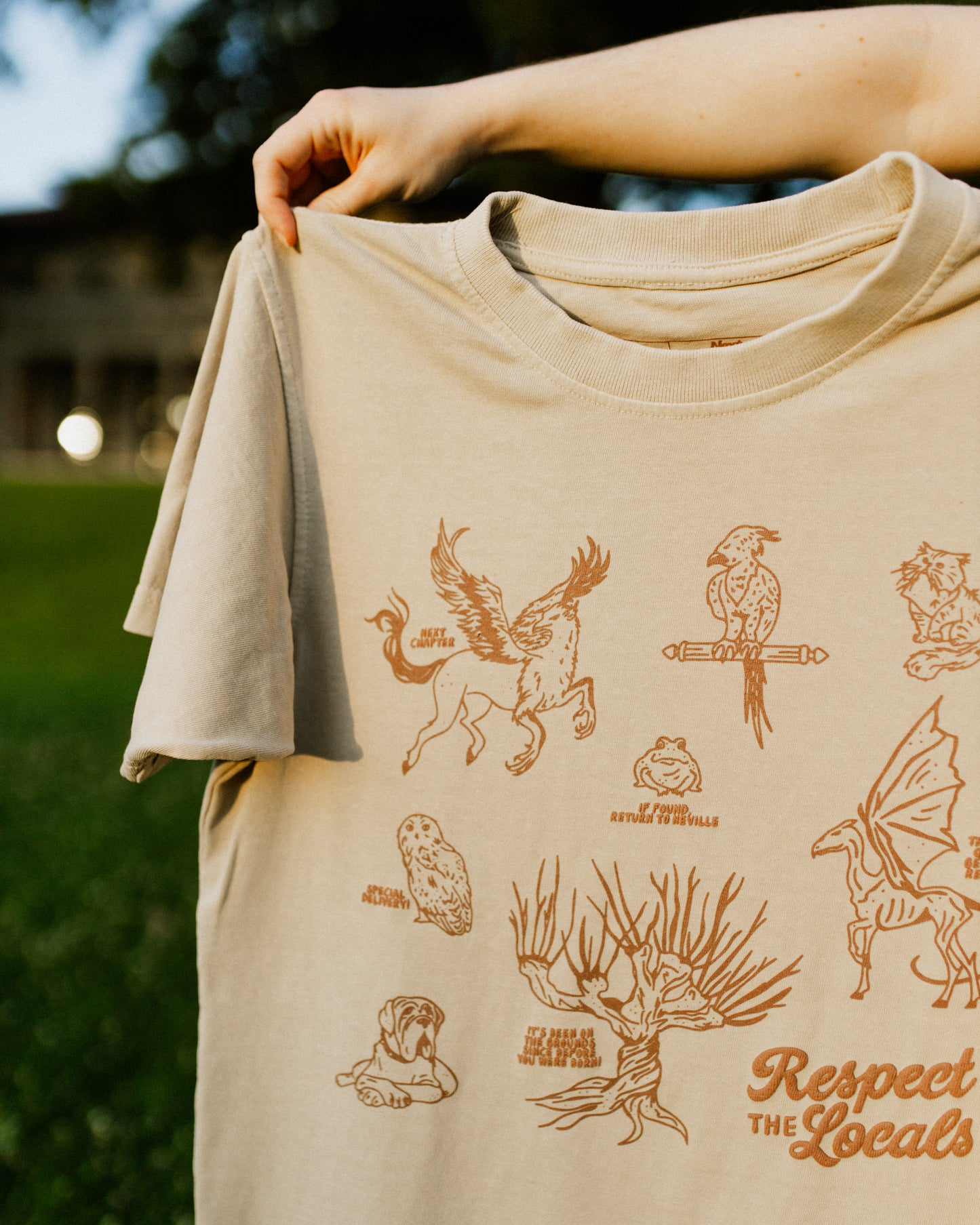 Beige t-shirt with brown illustrations and 'Respect The Locals' text held by a person outdoors. The t-shirt design is inspired by the creatures in Harry Potter.