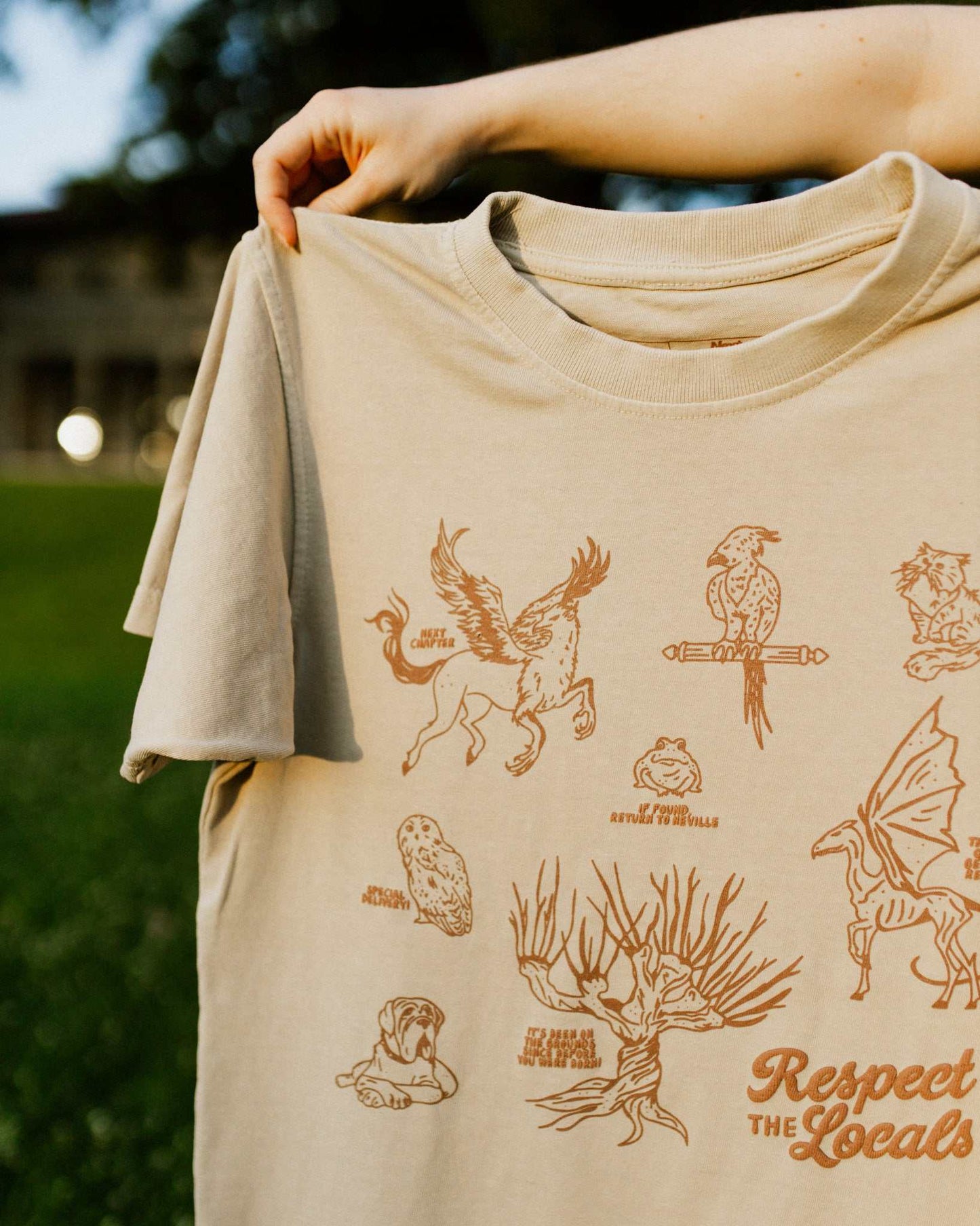 Beige t-shirt with brown illustrations and 'Respect The Locals' text held by a person outdoors. The t-shirt design is inspired by the creatures in Harry Potter.