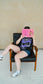 Person sitting on a chair holding a pink book with a plain background. She is wearing a black graphic tee with 'The Ourororos' design featuring an eye, mirror, and purple mist. Inspired by the A Court of Thorns of Roses book series by Sarah J. Maas.