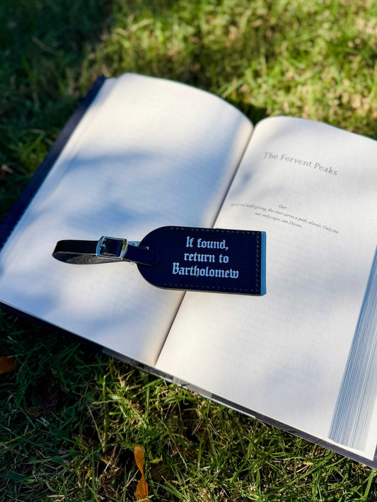 The Bartholomew luggage tags lie on a book outdoors. It reads "If found, return to Bartholomew" and is inspired by The Knight and the Moth by Rachel Gillig.