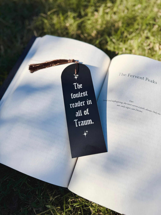 The Foulest Reader Bookmark lies on a book outdoors. It reads "The foulest reader in all of Traum" and is inspired by the book The Knight and the Moth by Rachel Gillig. The bookmark is black and white with a brown tassel attached.
