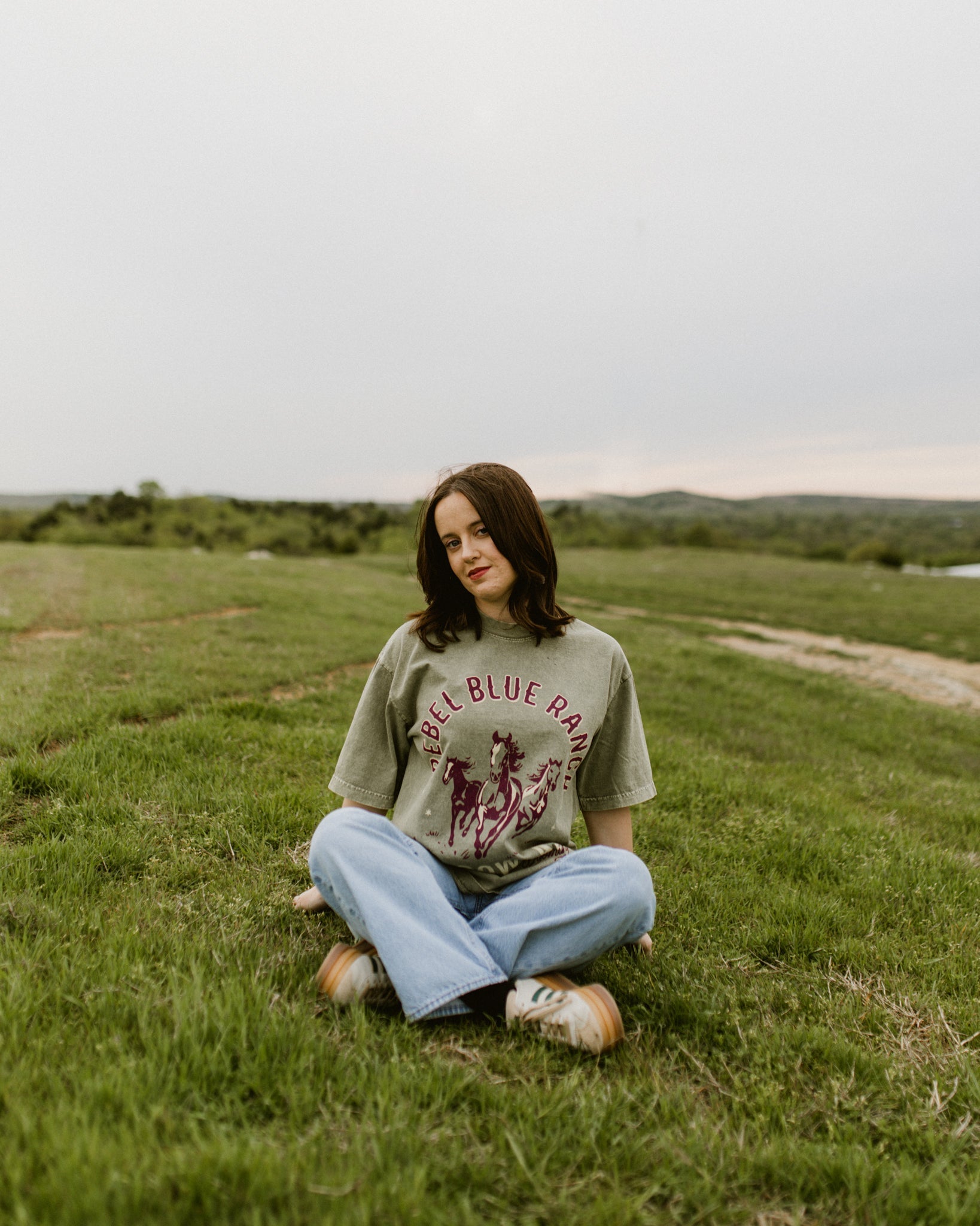 Rebel Blue Ranch Mustangs Tee. A woman wears a mineral wash olive green boxy oversized Rebel Blue Ranch tee with a horses design based on the cowboy romance book series by Lyla Sage that includes Done and Dusted, Swift and Saddled, Lost and Lassoed, and Wild and Wrangled. Meadowlark bookish merch. Lyla Sage merch.