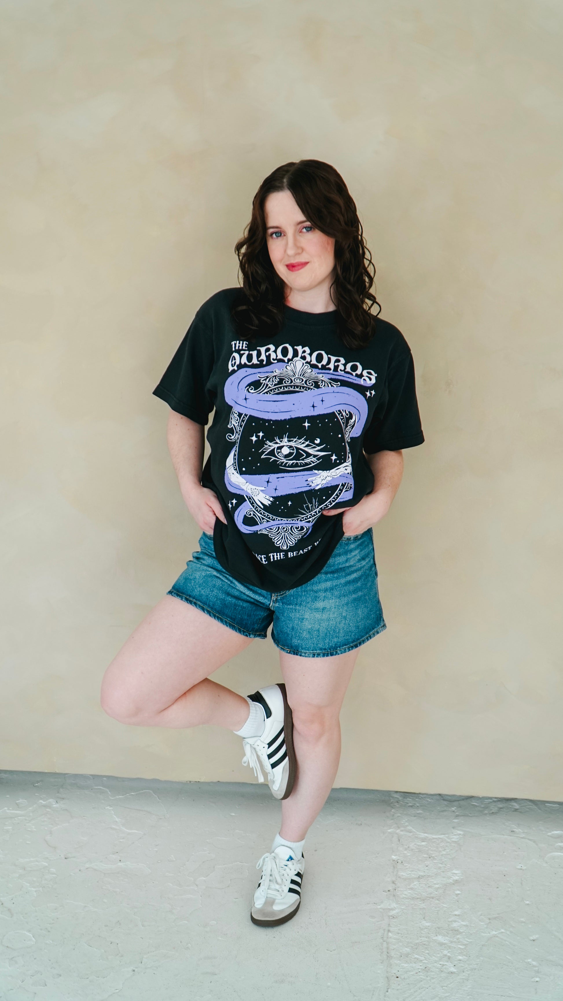 Person wearing a black graphic t-shirt and denim shorts against a beige wall. She is wearing a black graphic tee with 'The Ourororos' design featuring an eye, mirror, and purple mist. Inspired by the A Court of Thorns of Roses book series by Sarah J. Maas.