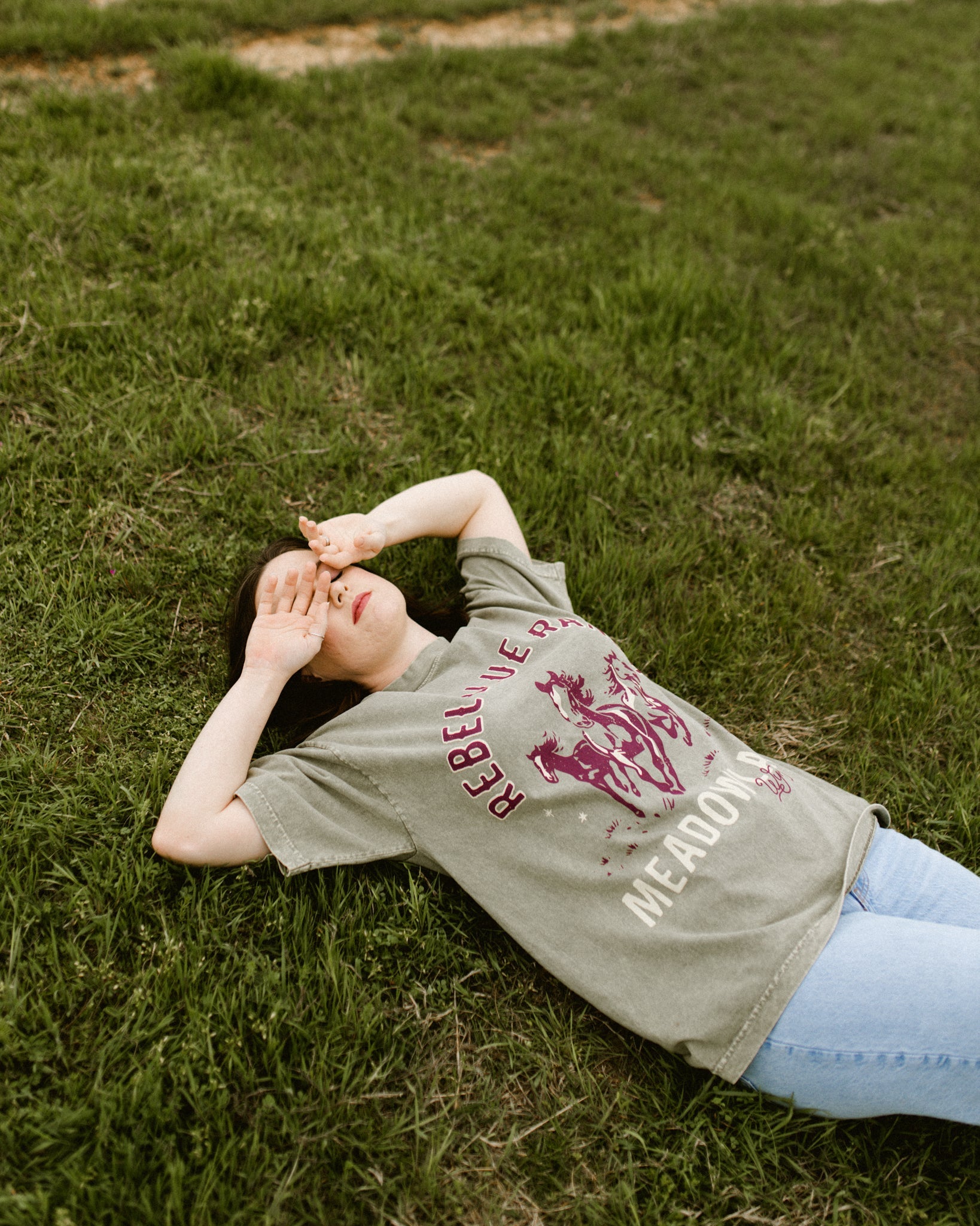 Rebel Blue Ranch Mustangs Tee. A woman wears a mineral wash olive green boxy oversized Rebel Blue Ranch tee with a horses design based on the cowboy romance book series by Lyla Sage that includes Done and Dusted, Swift and Saddled, Lost and Lassoed, and Wild and Wrangled. Meadowlark bookish merch. Lyla Sage merch.